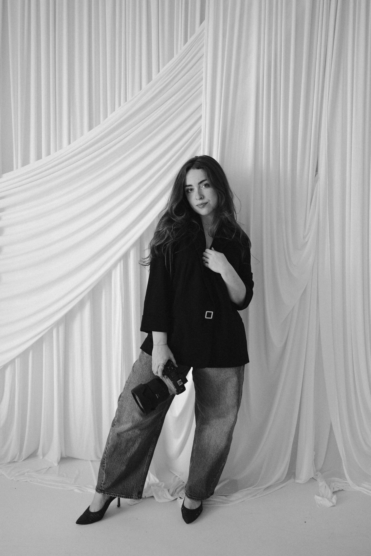 Black and white photo of a woman in a black blazer and jeans holding a bag, posing in front of white draped curtains.