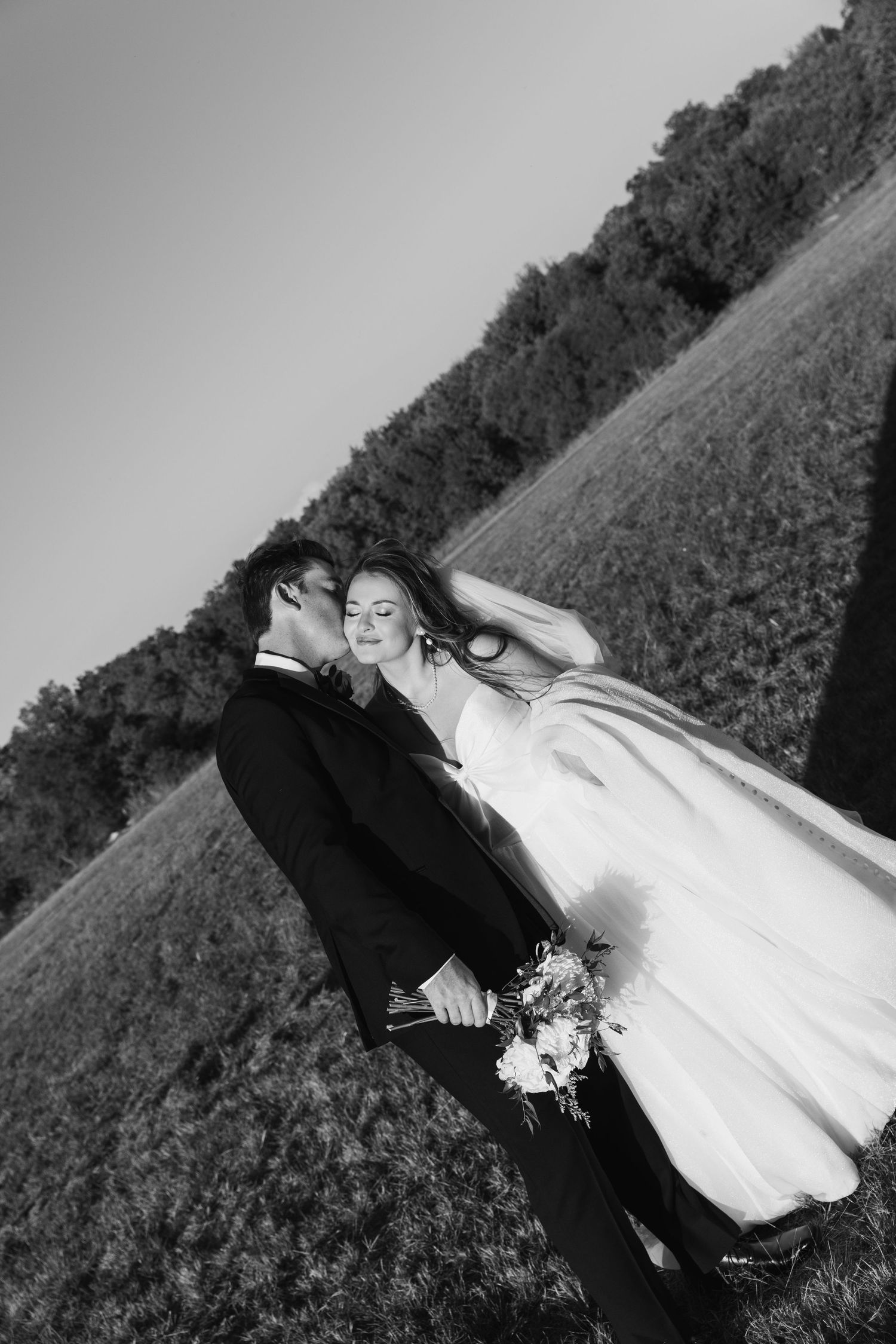 A black and white wedding photo shows a couple embracing on a grassy hillside in an artistic tilted angle composition.