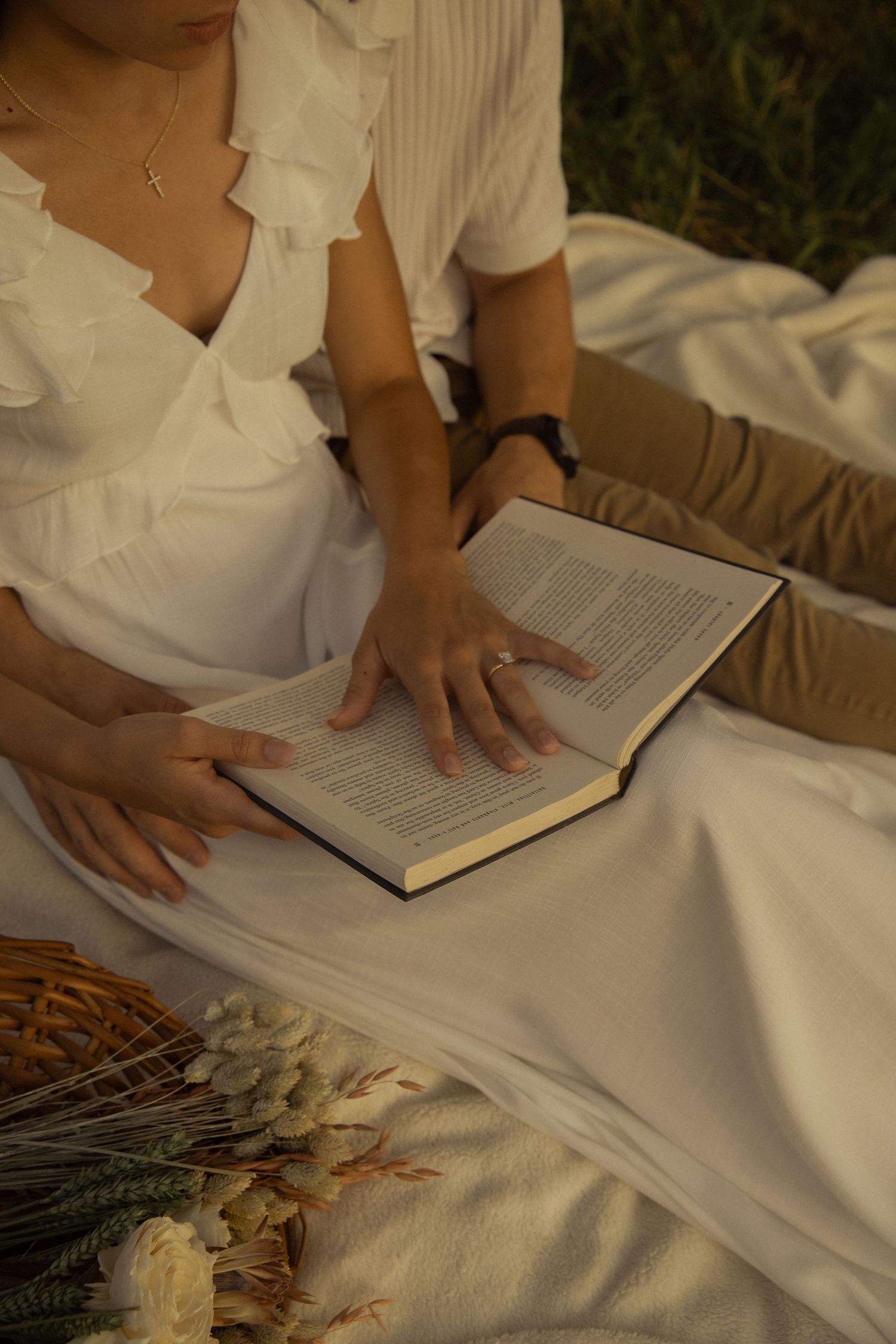 Person in white clothing reading a book while lying on a white blanket outdoors.