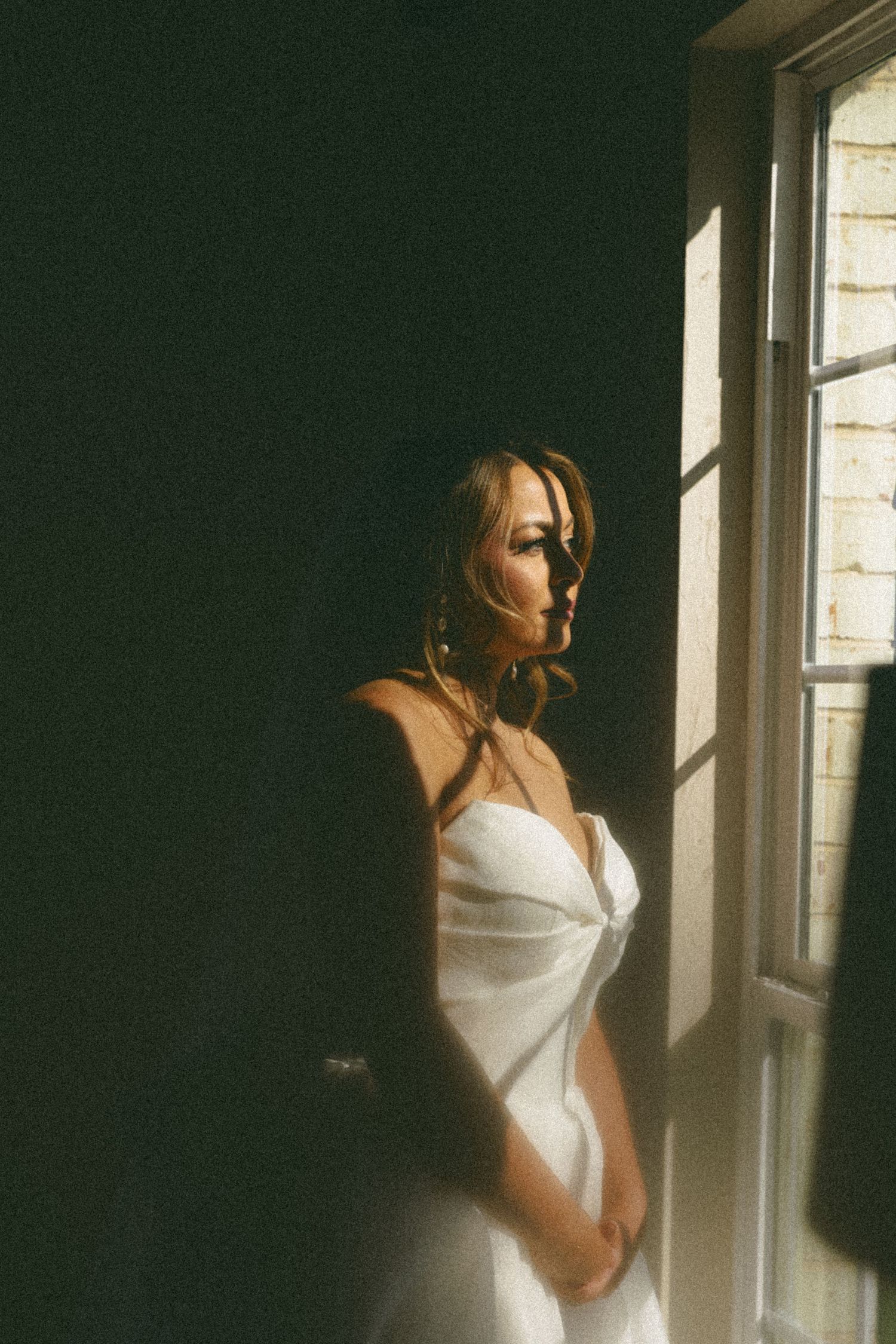 A figure in a white dress stands by a sunlit window creating a moody, vintage-style portrait with dramatic lighting.