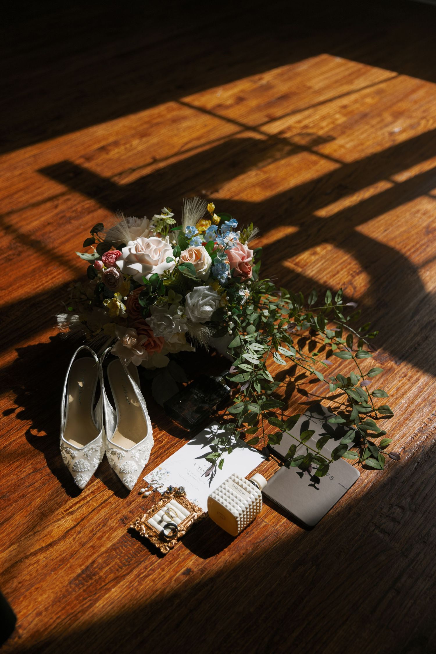 Wedding accessories including lace shoes, bouquet, and jewelry arranged on a sunlit wooden floor with dramatic shadows.