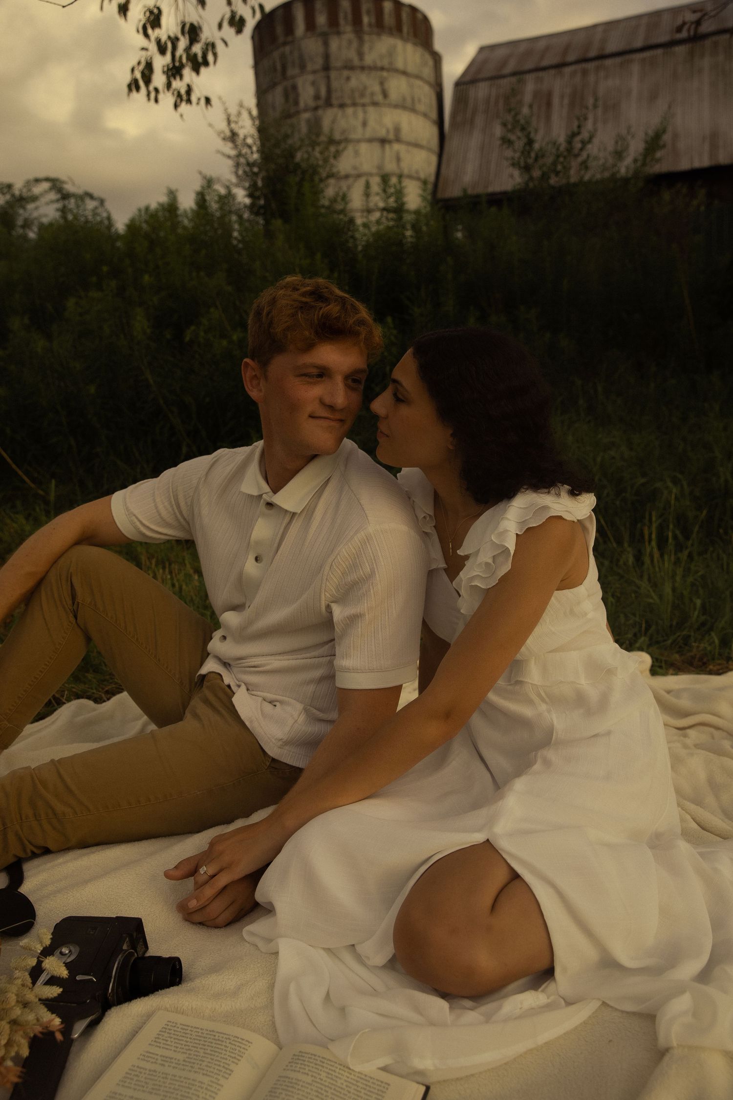 A romantic picnic scene with books and blankets near old farm buildings.
