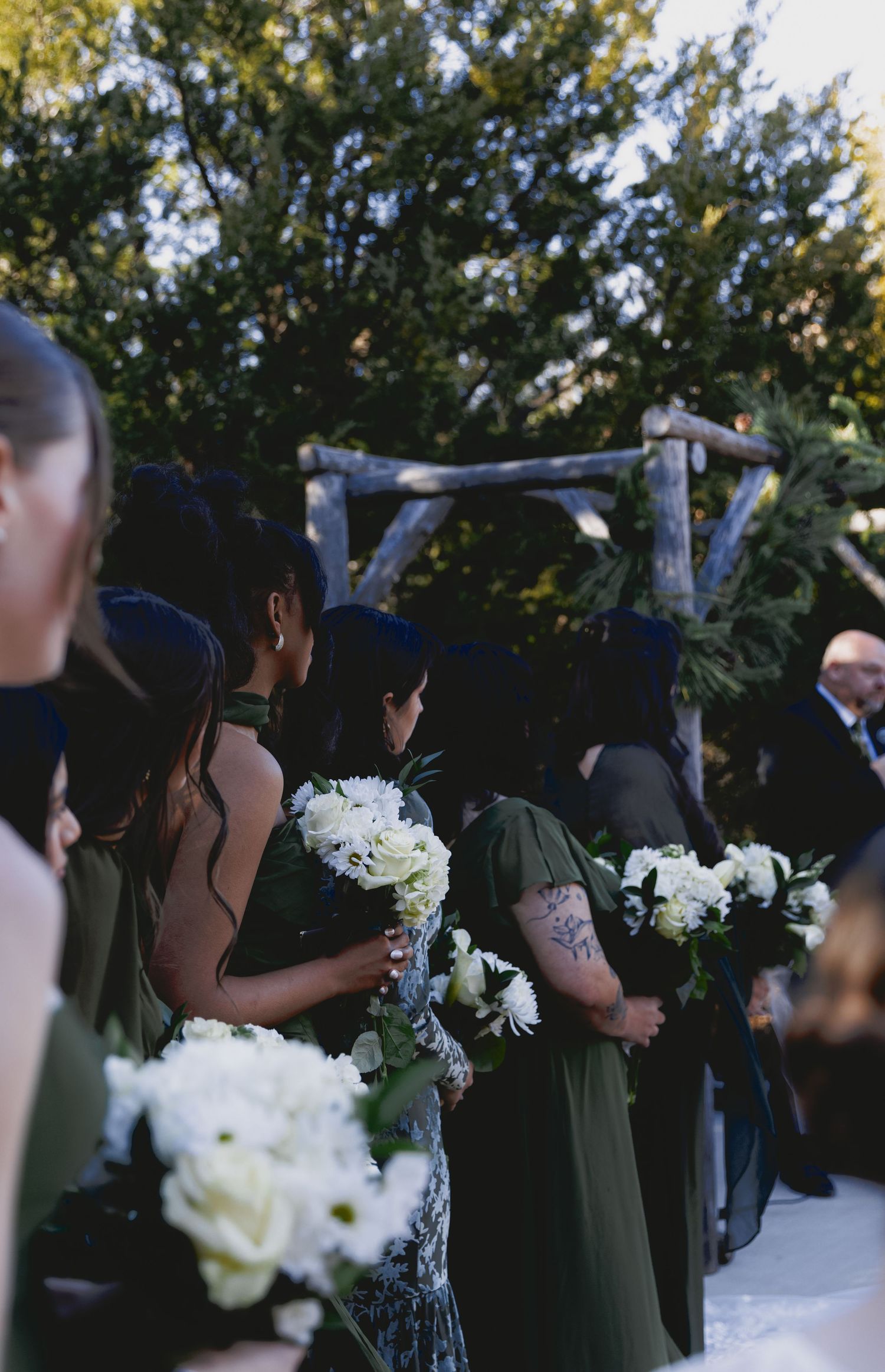 Bridesmaids in dark dresses hold white floral bouquets during an outdoor wedding ceremony under a rustic wooden arch.