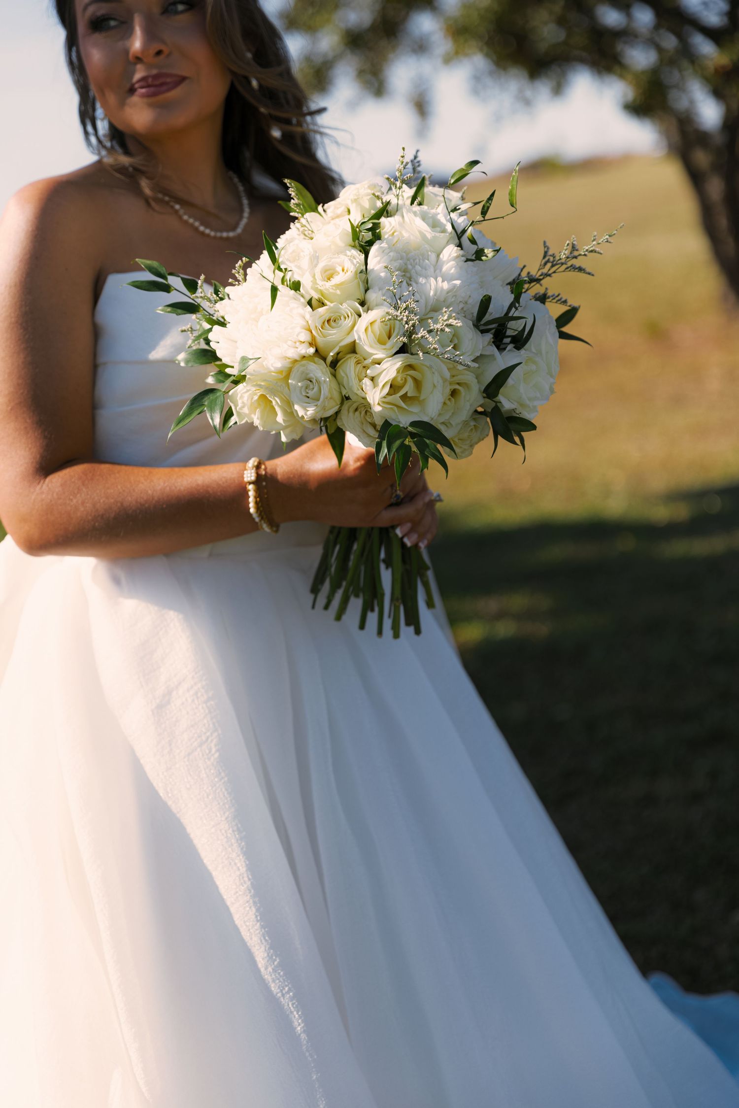 A bride in a white strapless gown holds an elegant bouquet of white roses and greenery on her wedding day.