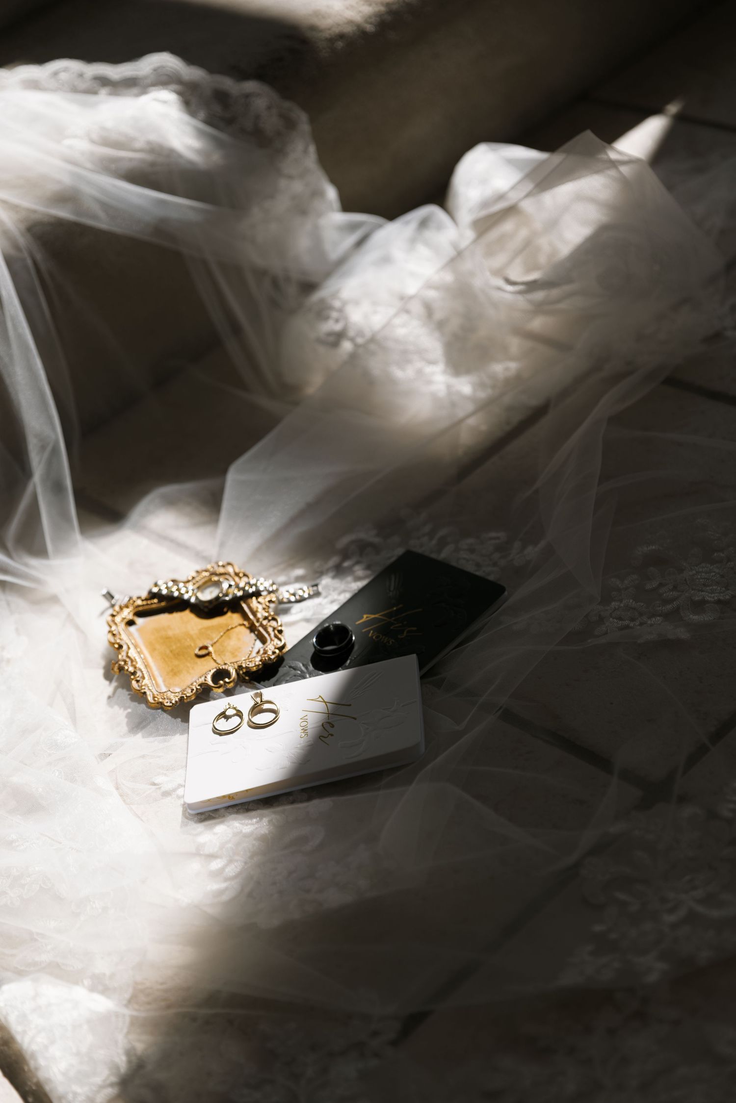 A golden wedding ring and engagement ring rest on delicate white tulle fabric against a dark background.