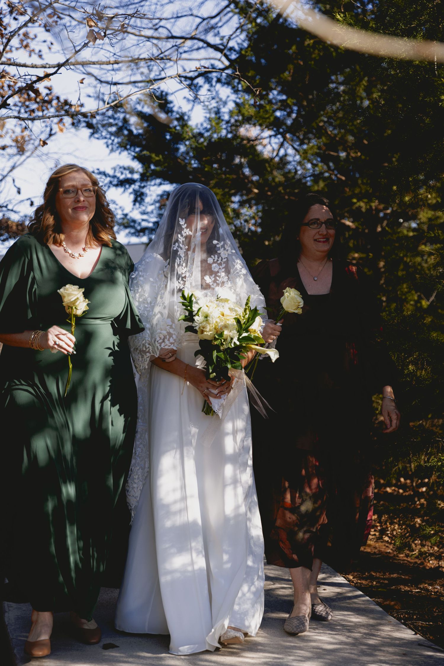 Wedding couple stands together in dappled sunlight holding bouquets during outdoor autumn ceremony.