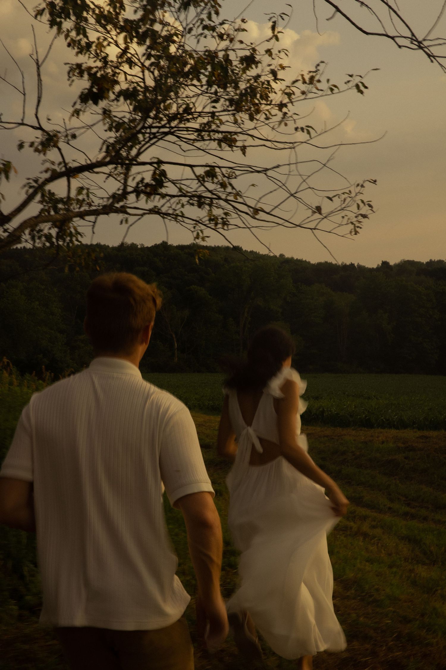A couple in white clothing walking together through a rural landscape at sunset.