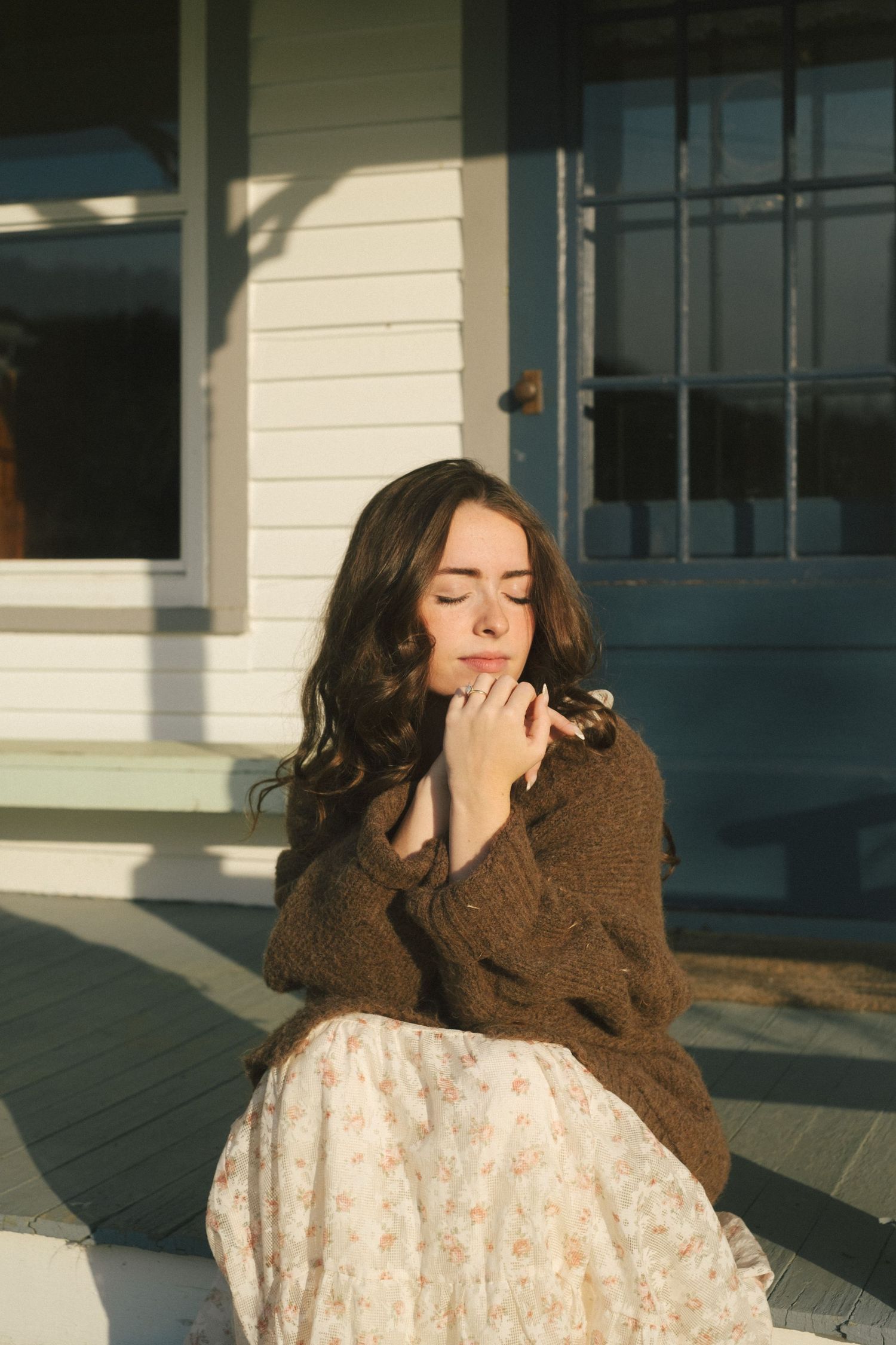 A person in a brown sweater and white lace dress poses against a modern building exterior in warm sunlight.