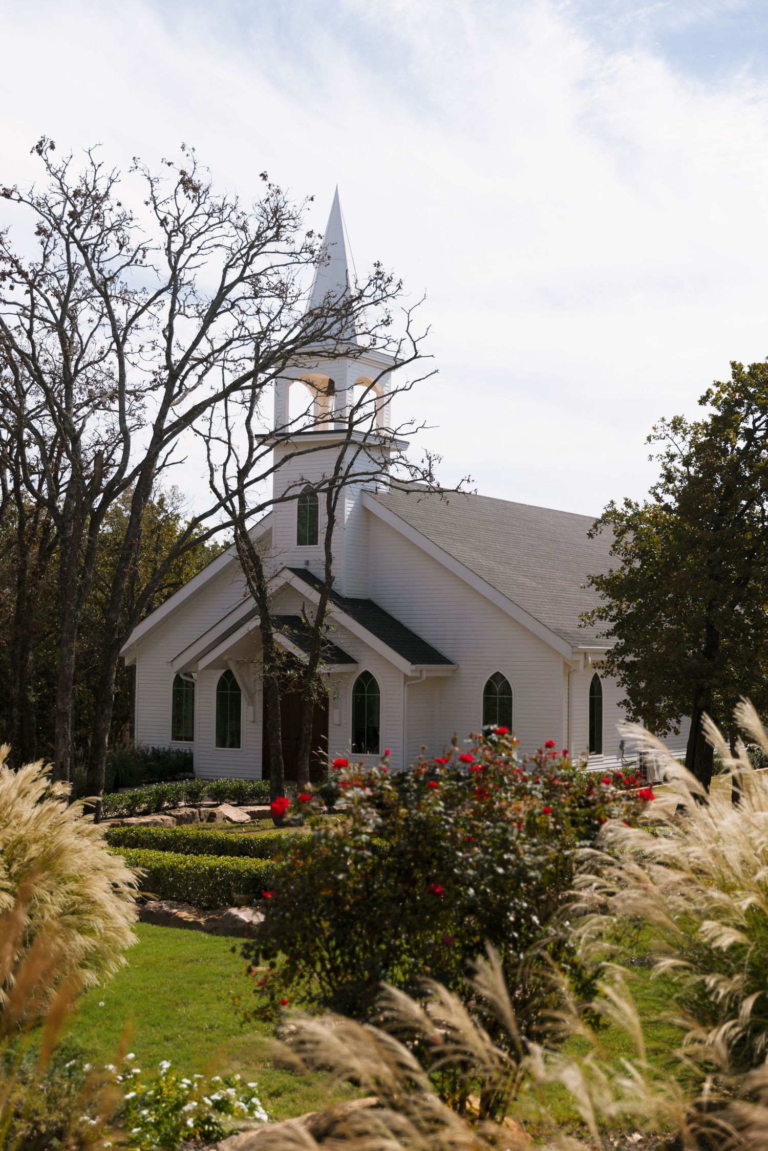 A white country church with a steeple sits among ornamental grasses and flowering bushes on a cloudy day.