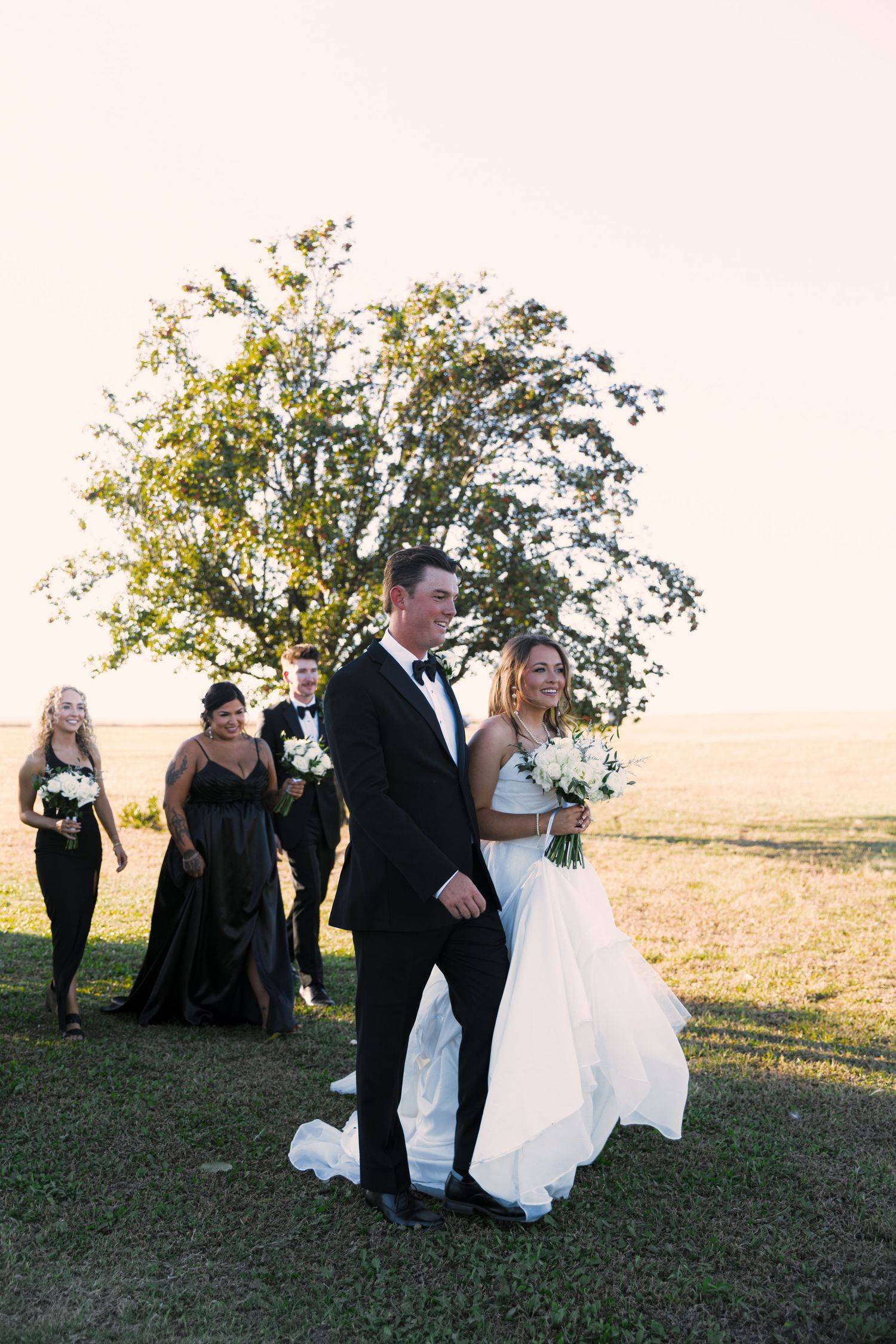 Wedding party in black attire walks through a grassy hilltop field at sunset during an outdoor celebration.