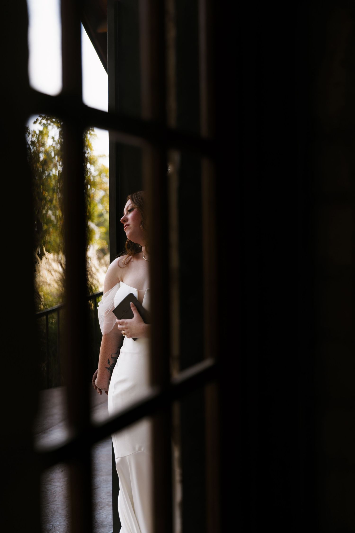 A figure in a white wedding dress stands in dramatic silhouette against a bright window in an elegant interior setting.
