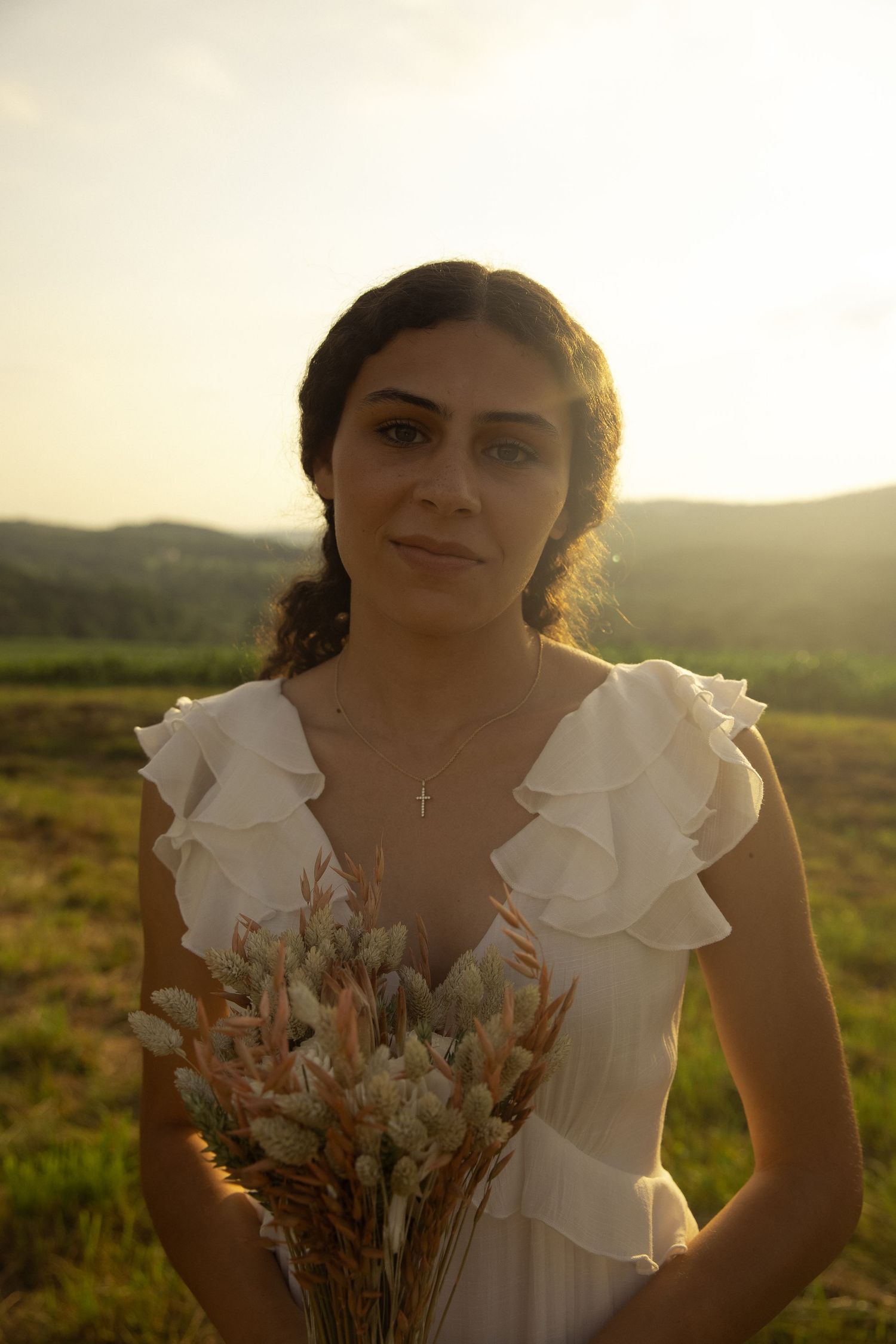 Person in white ruffled dress holding dried flowers in golden sunlight.