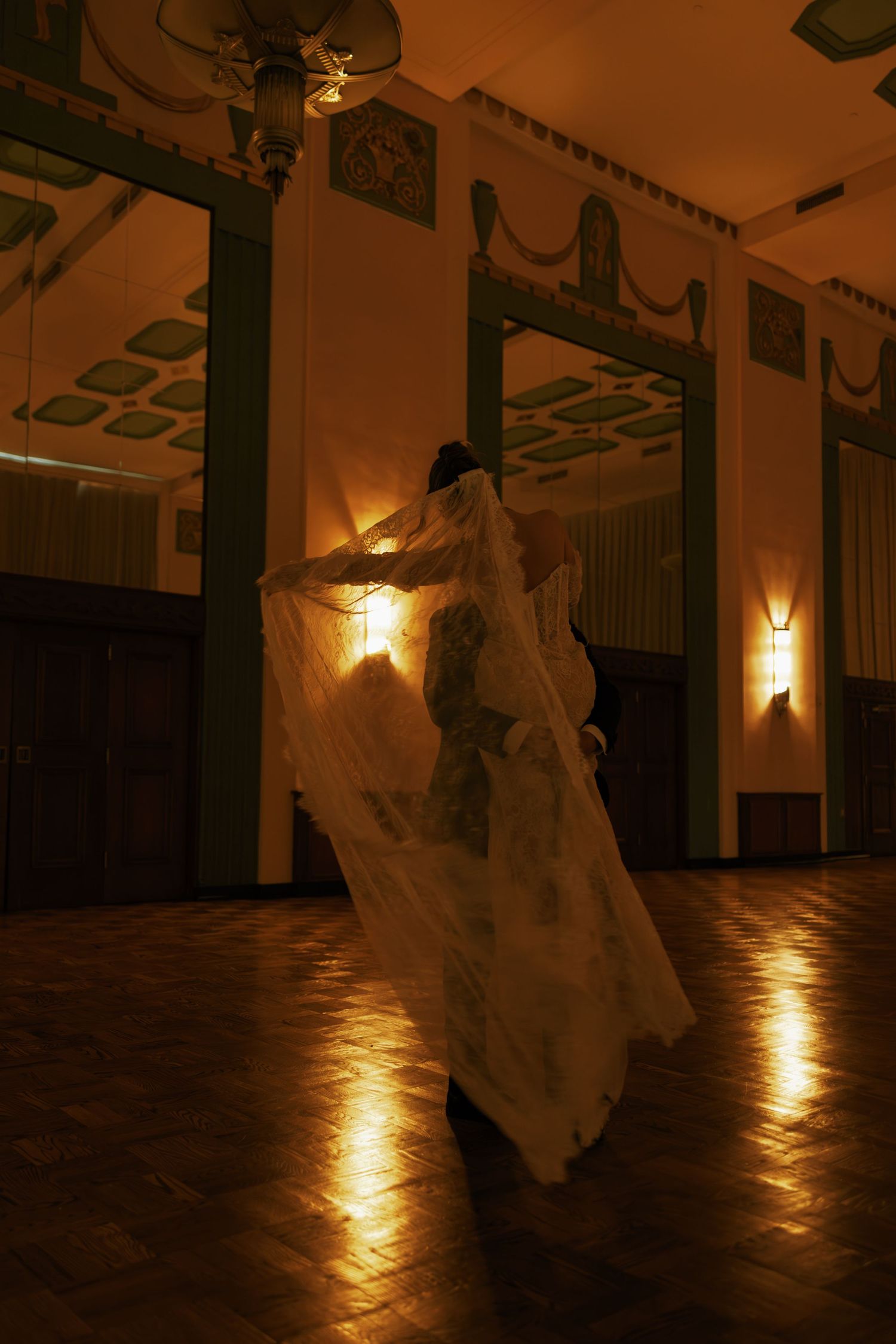 Bride in flowing white wedding gown and veil dancing alone in an elegant warmly lit ballroom with parquet floors.