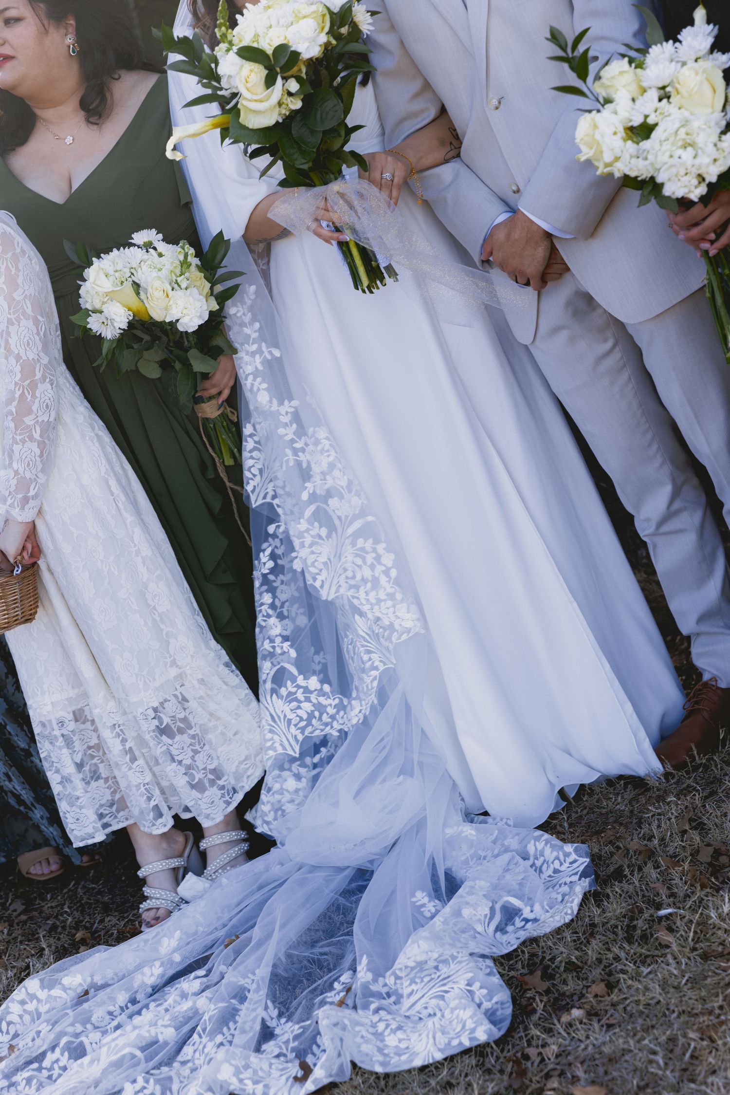 Close-up view of white lace wedding dress hem and light gray suits at an outdoor ceremony.