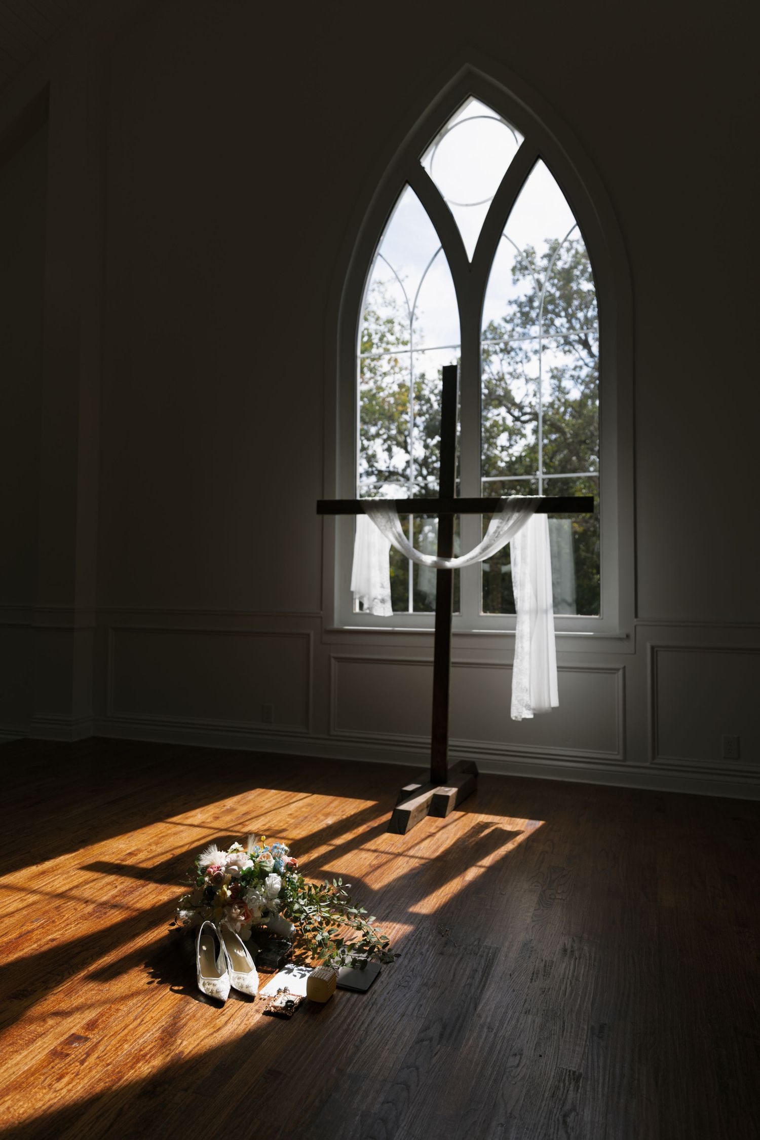 Sunlight streams through a gothic church window onto a wooden floor where a wedding dress and flowers are arranged.