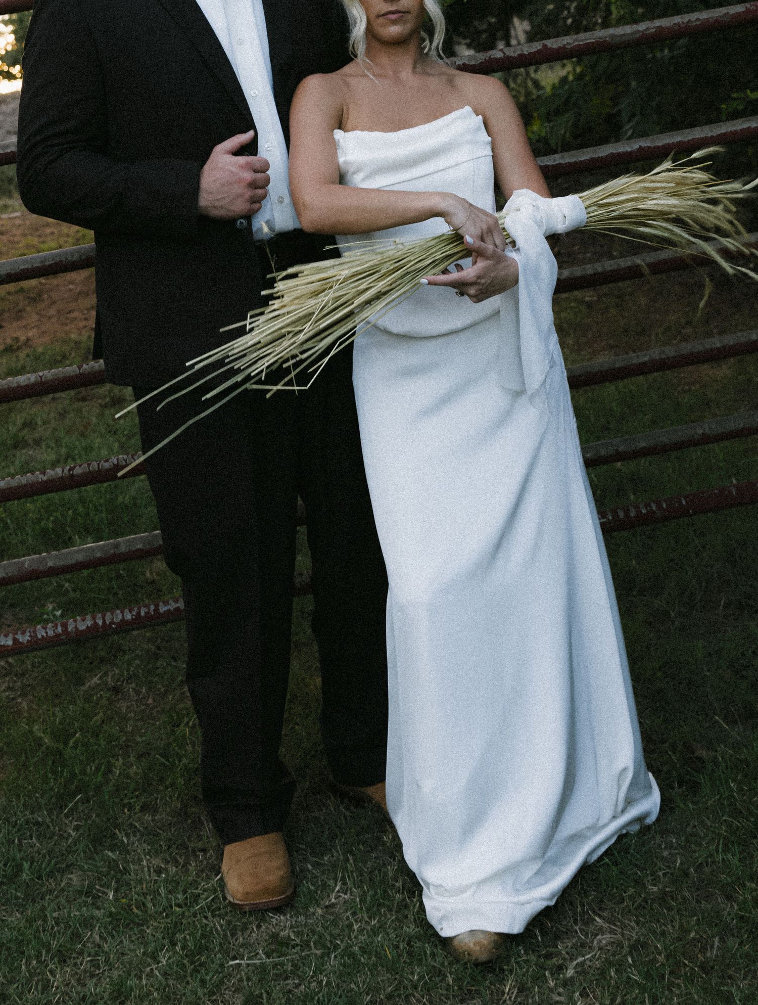 Couple in formal wedding attire holding wheat stalks against rustic fence.
