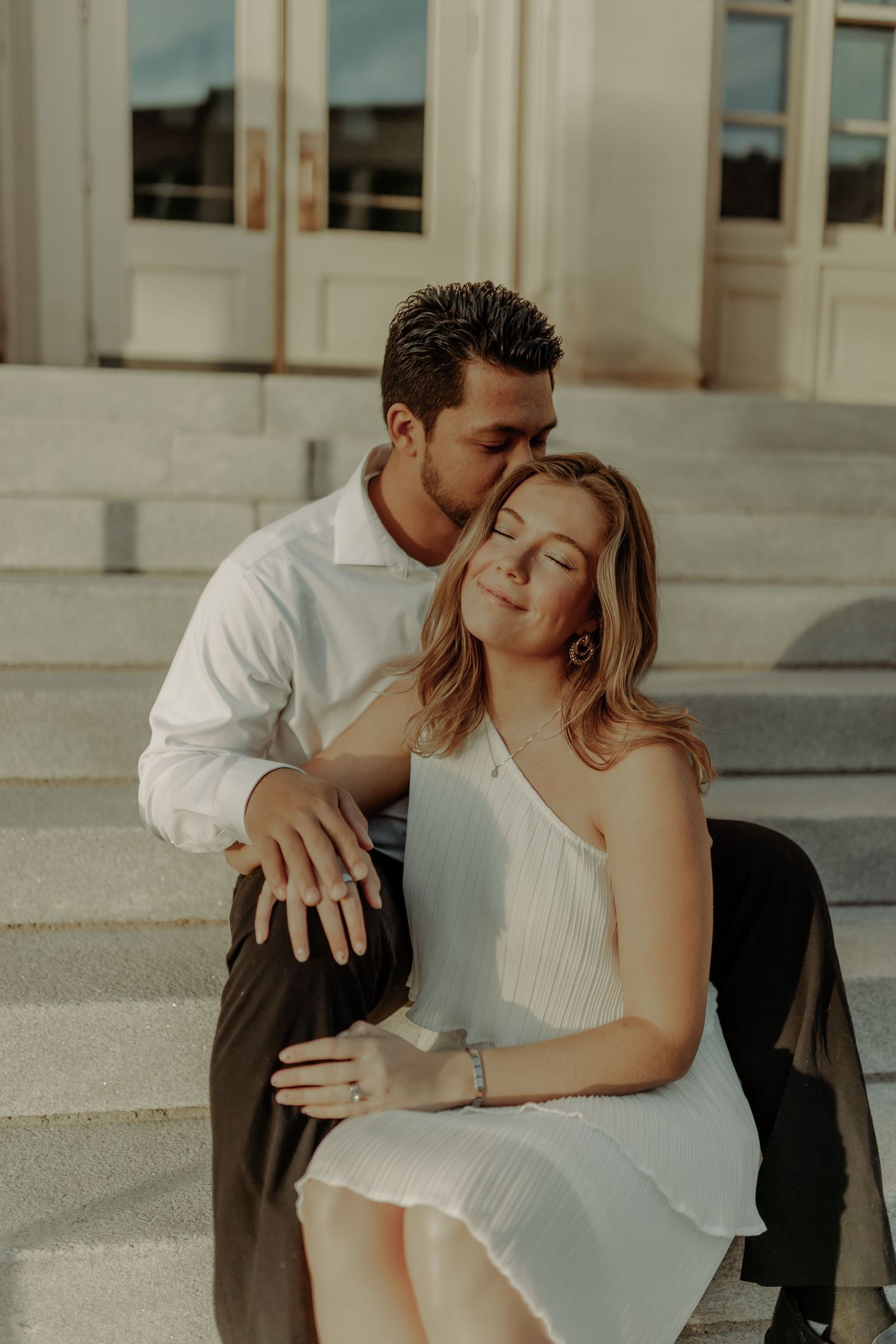 downtown grand junction couple sitting on steps of courthouse smiling and he is kissing her cheek during engagement photoshoot