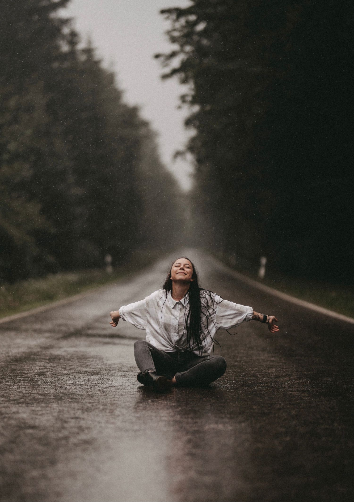 Person sitting cross-legged with arms outstretched on a rainy forest path.