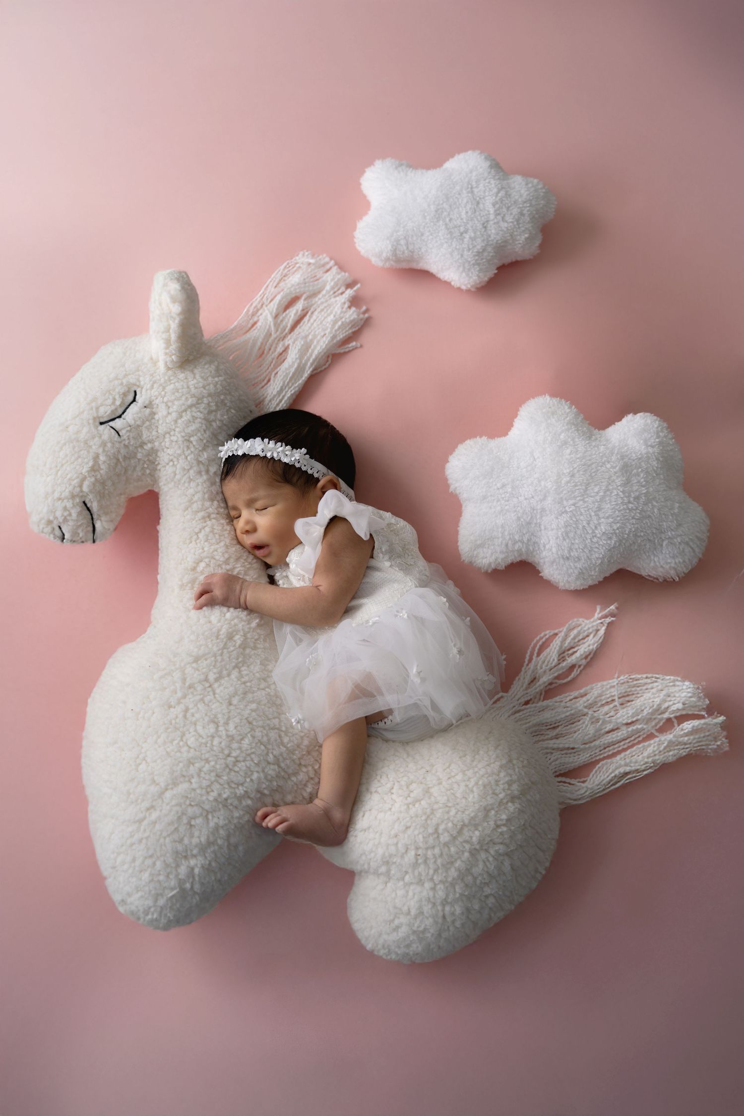 A sleeping baby nestled among fluffy white cloud-shaped pillows and delicate lace on a pink background.