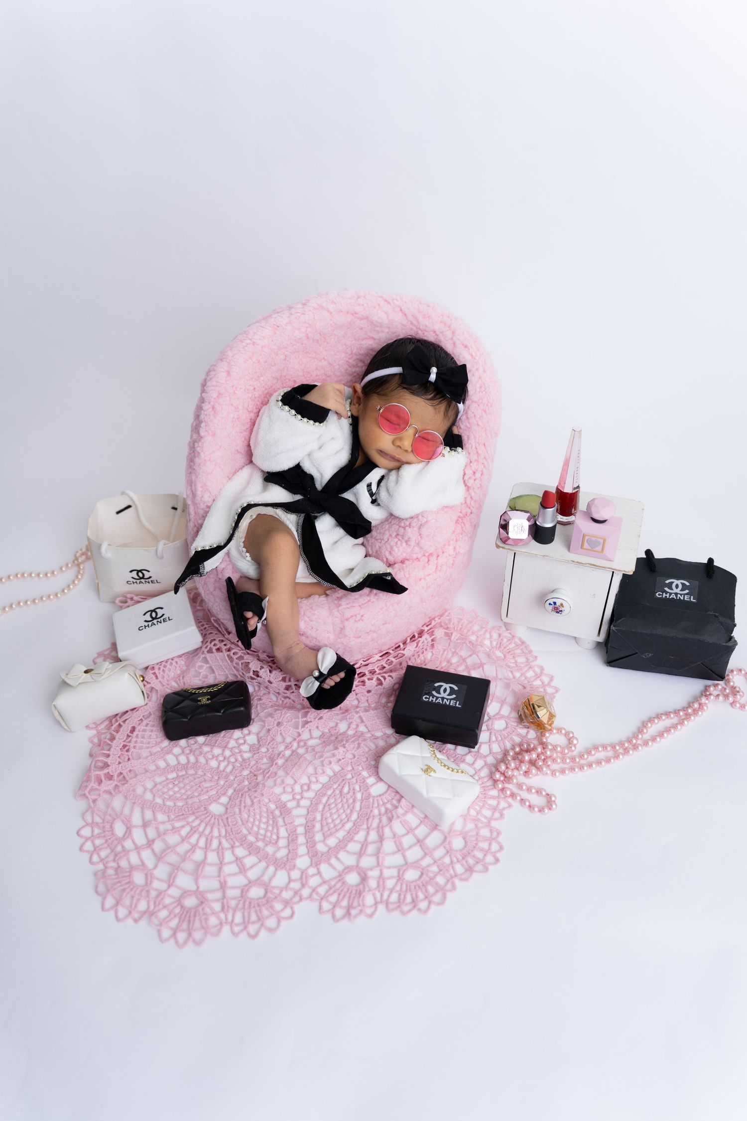 Luxury fashion items laid out on a pink doily with black boxes, white flowers and pearls creating an elegant flat lay.