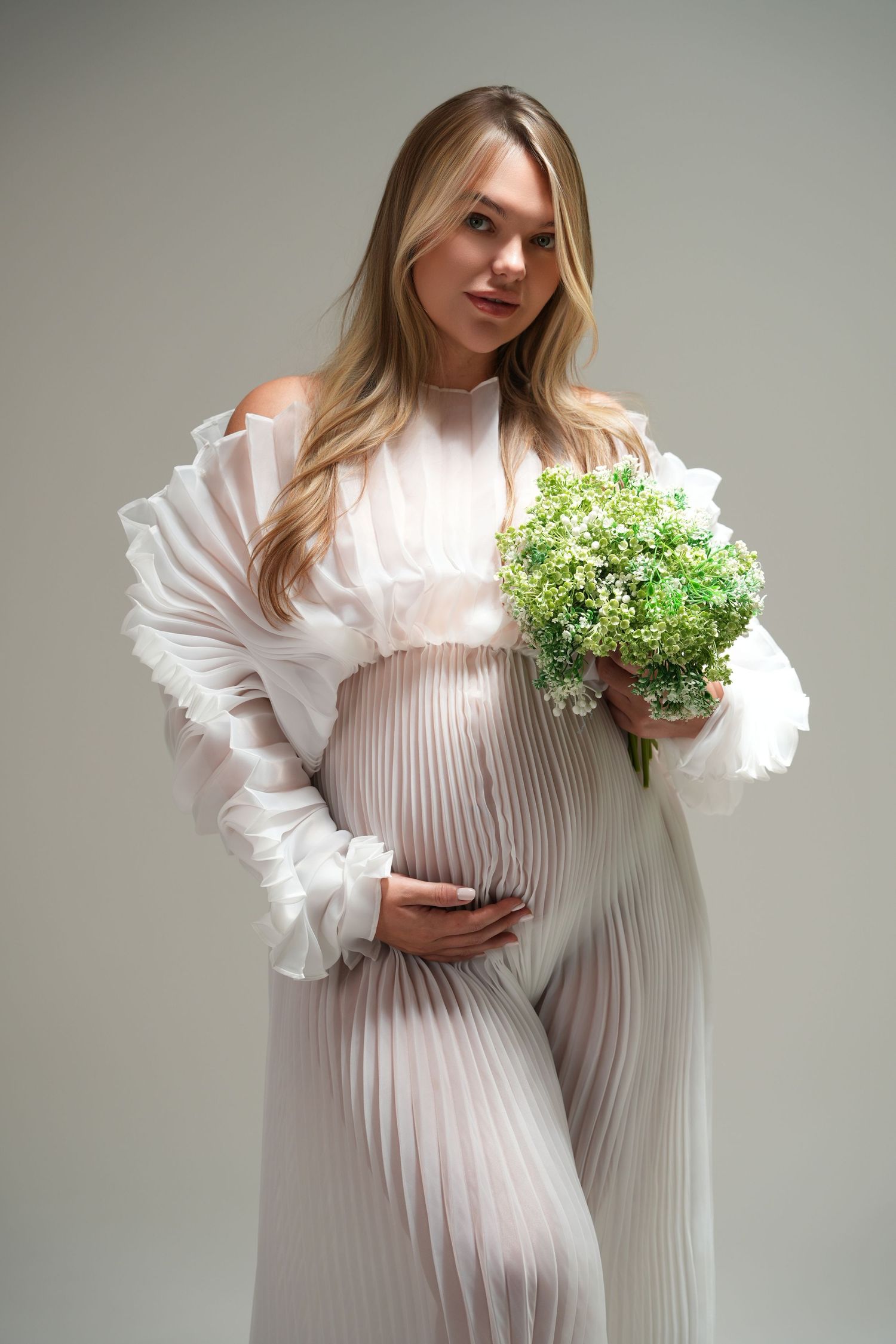 Ethereal figure in pleated white dress holds fresh green flowers against neutral backdrop in artistic portrait series.