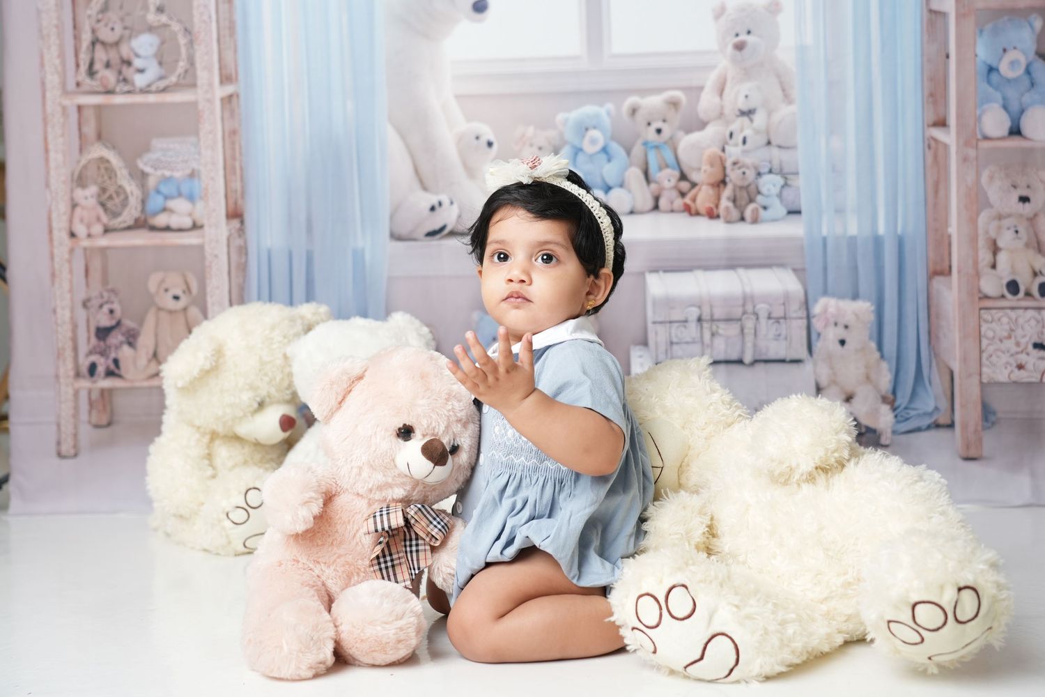 A baby in white onesie sits among plush teddy bears and soft toys in a dreamy pastel nursery setting.