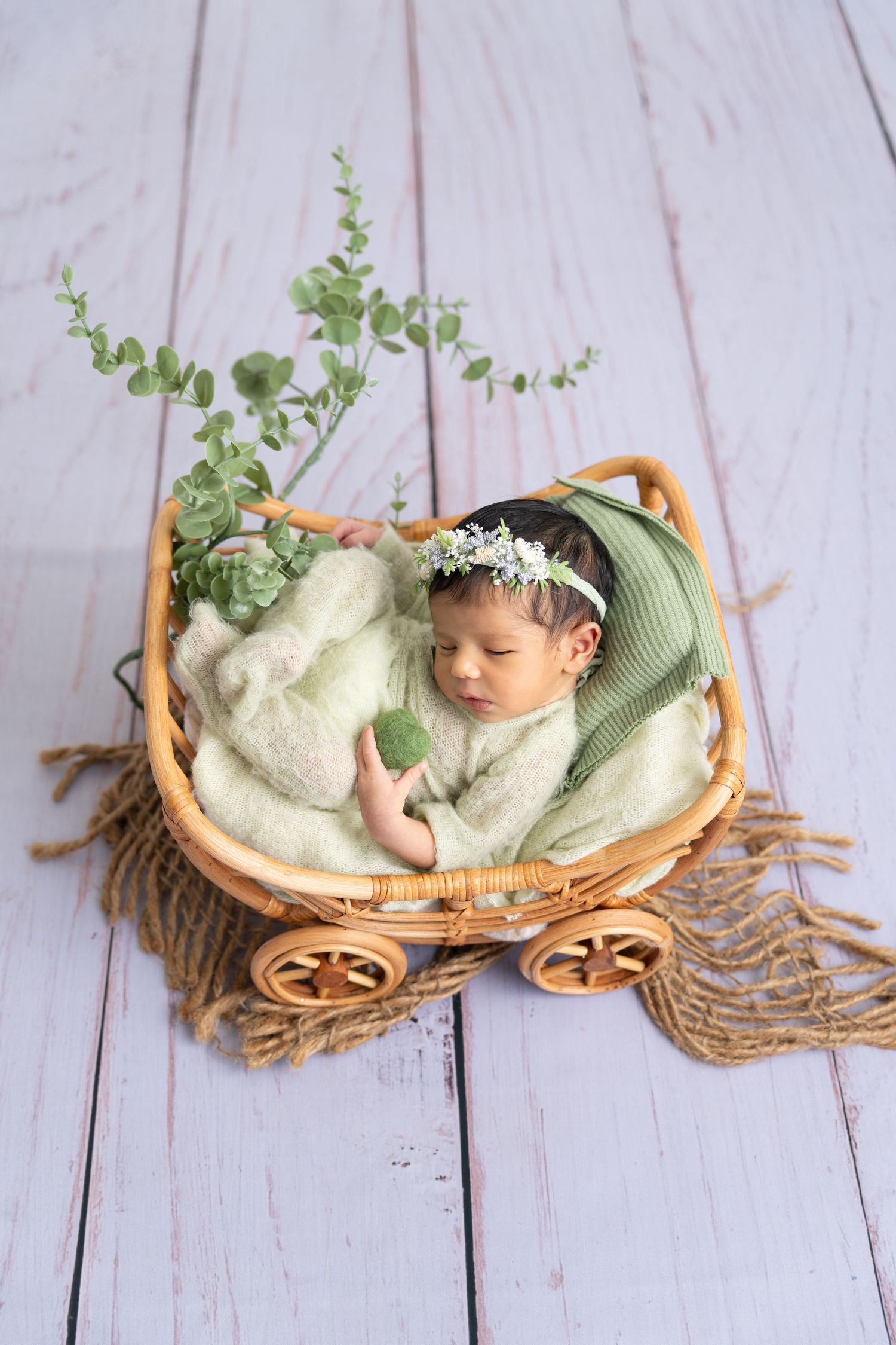 A sleeping baby in a green wrap rests in a wooden wagon decorated with greenery on a white wooden floor.