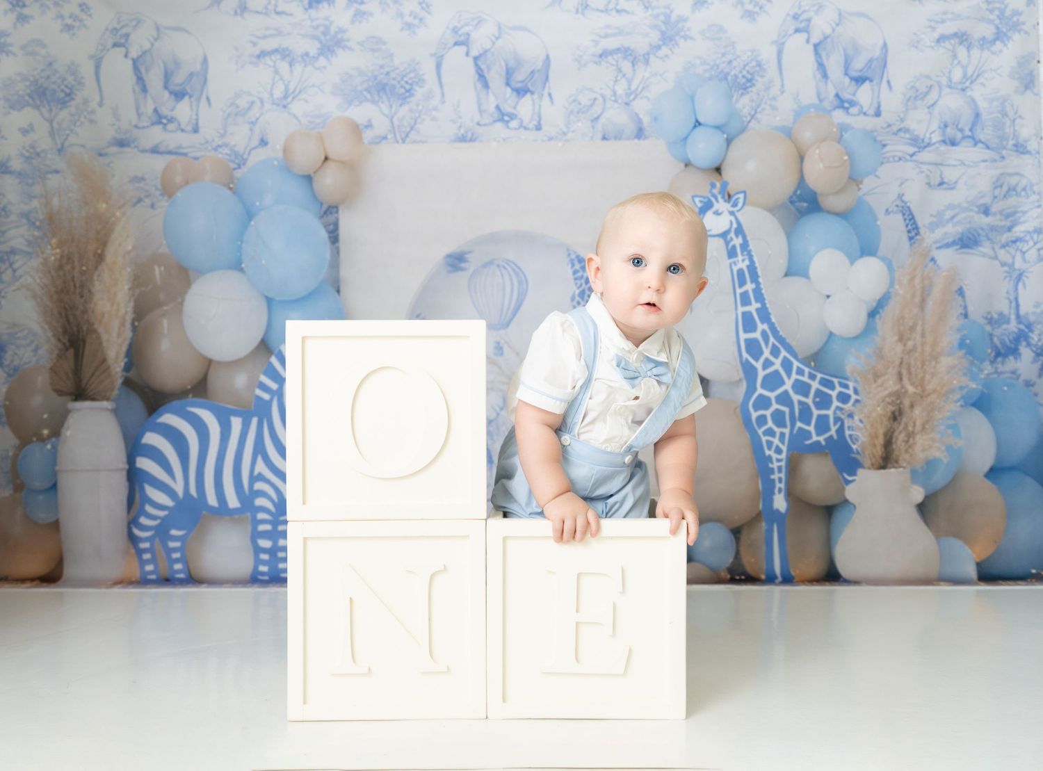 Baby in white onesie sits on white decorative blocks with blue safari-themed backdrop and balloons.