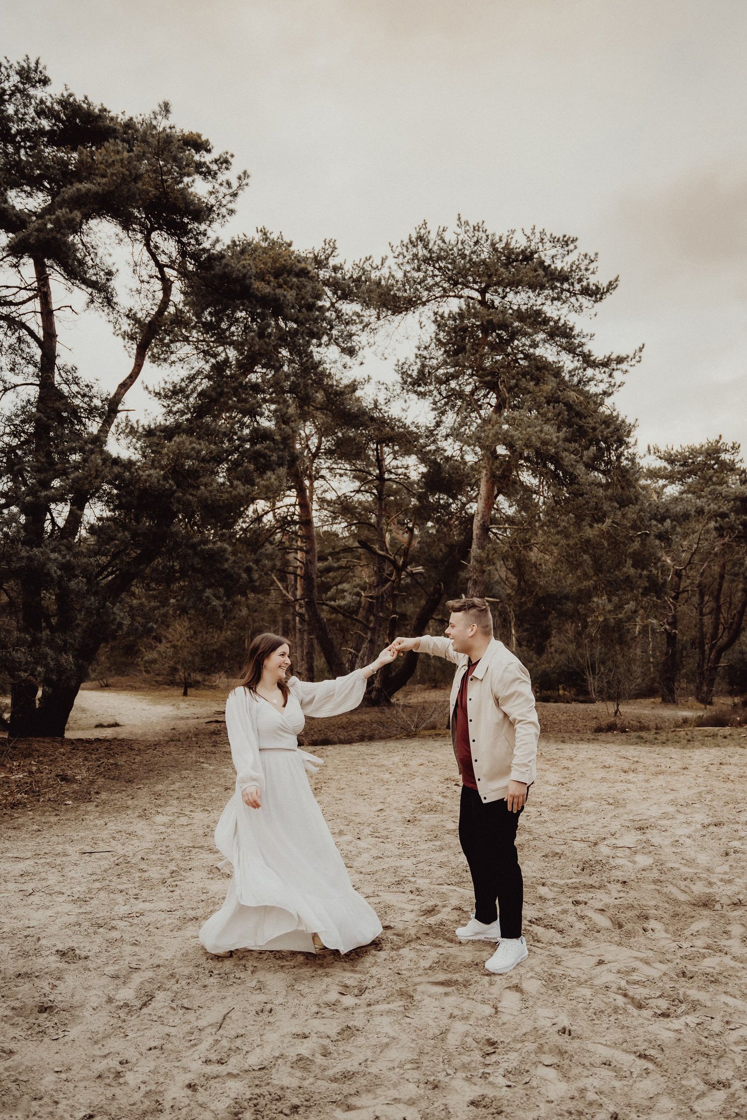 A couple in formal attire dances together in a rustic outdoor setting with pine trees in the background.