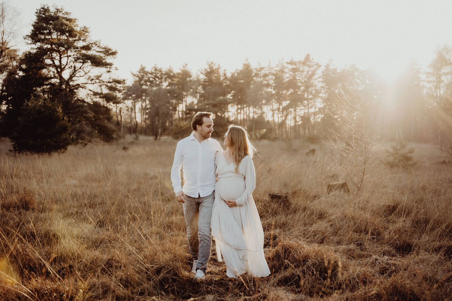 A couple poses together in a golden sunlit field wearing light colored clothing.