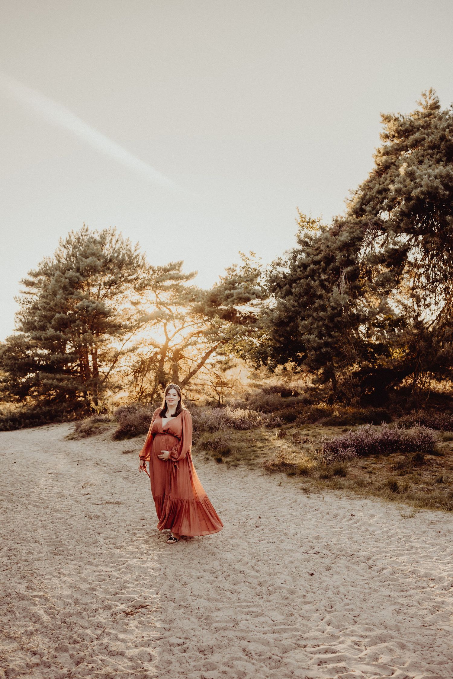 Pregnant woman in flowing rust-colored dress walking along sandy path in nature during golden hour.