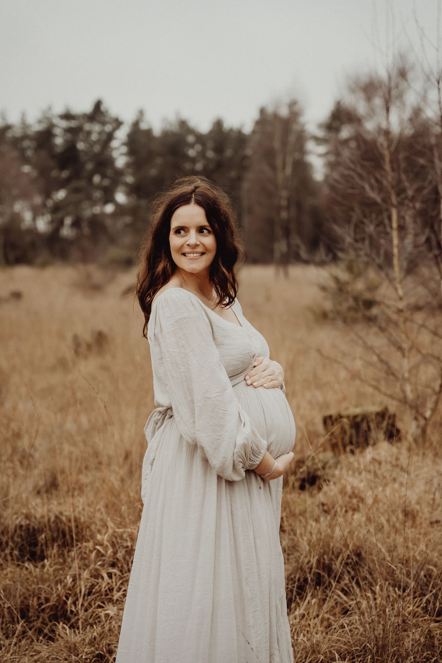 A woman in a flowing white dress cradles her pregnant belly in a sunlit field.