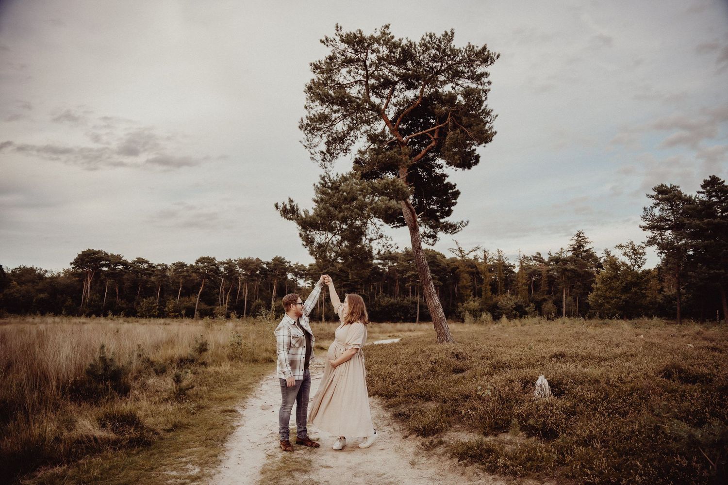 A couple stands together on a dirt path in a scenic meadow with tall trees.