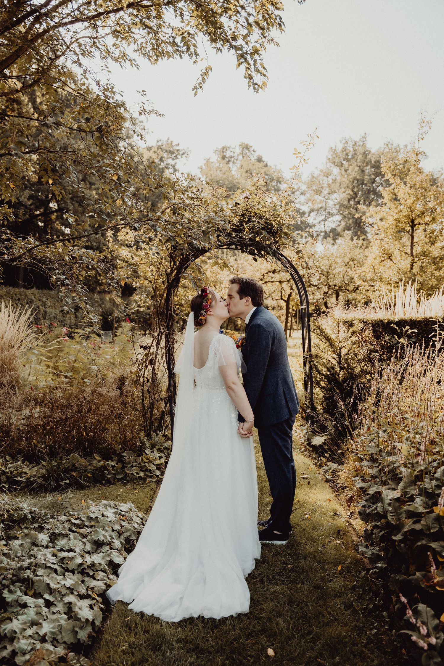 Newlyweds stand under a rustic garden arch surrounded by autumn foliage.