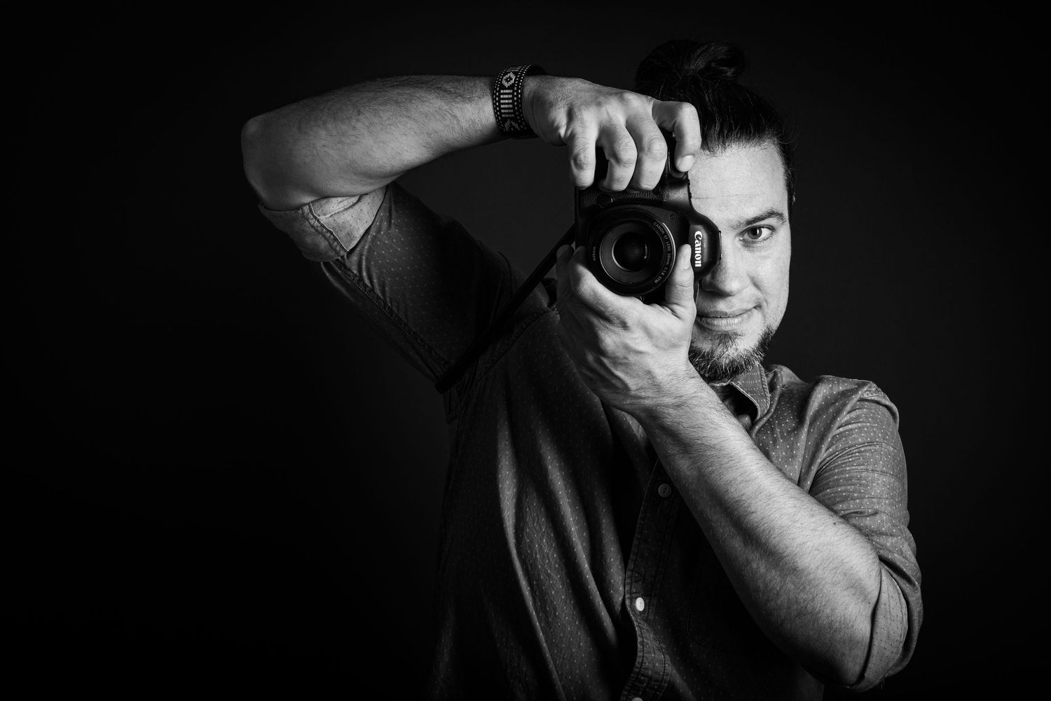 Black and white portrait of a photographer holding a DSLR camera up to their eye against a dark background.