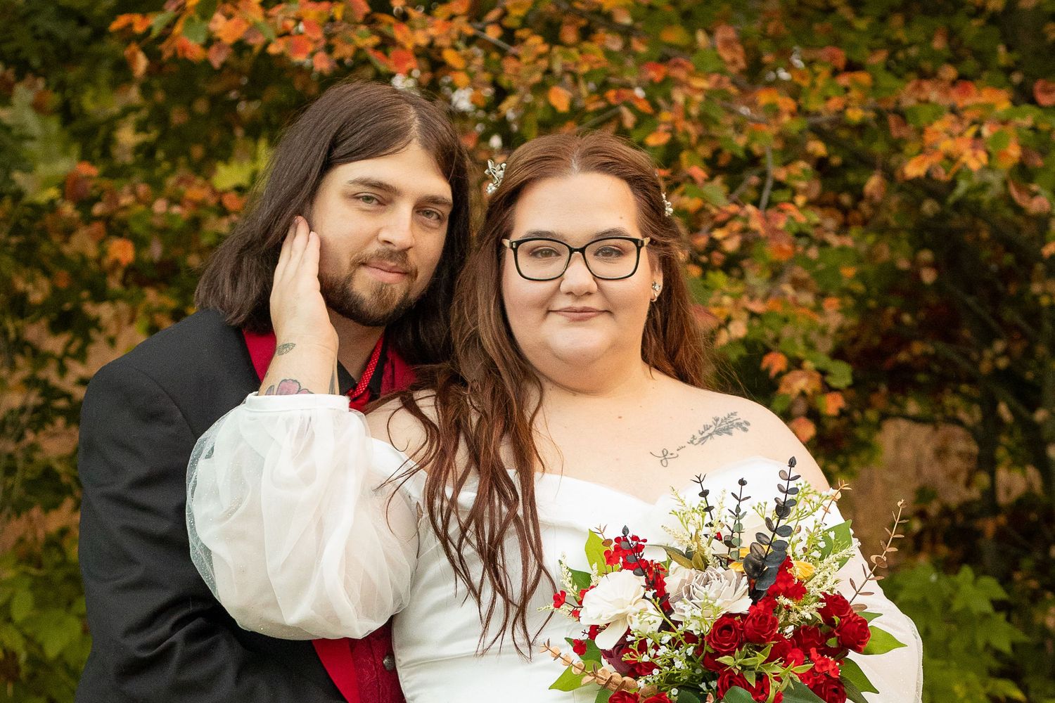 A couple poses together on their autumn wedding day with a red and white bouquet and fall foliage in the background.