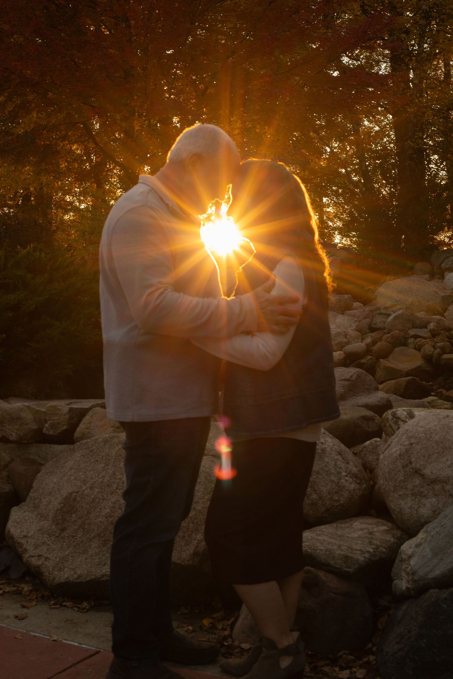 Two figures stand in silhouette against a dramatic sunset with sun rays bursting through trees and rocky terrain.