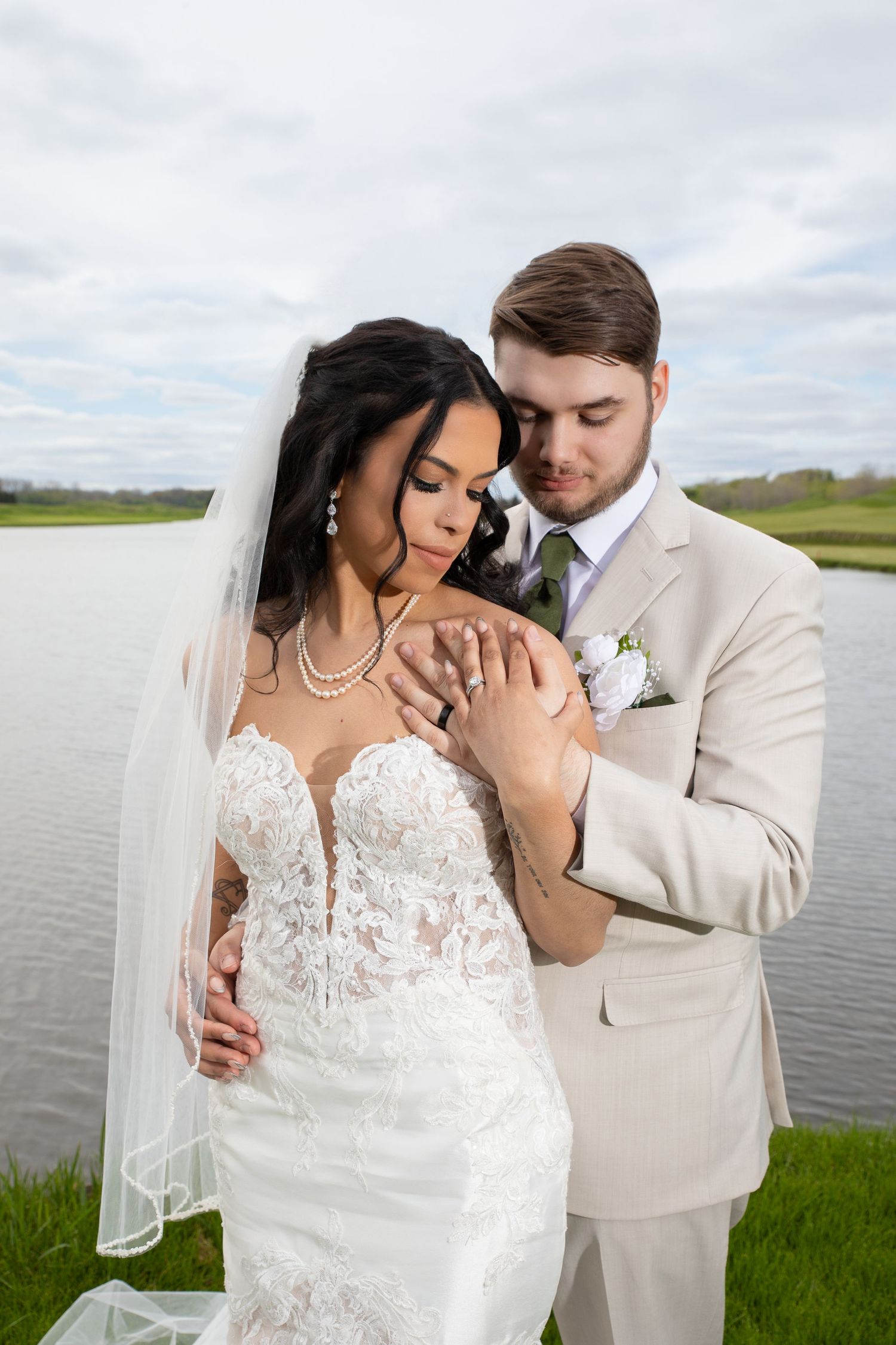 Couple embraces by a lake during wedding photos, bride wearing lace dress and veil with groom in beige suit.