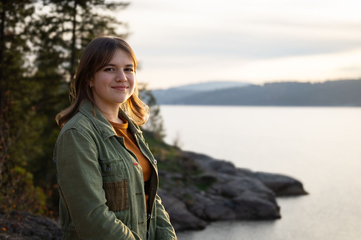 A smiling young woman stands before a scenic lake and rocky shoreline at sunset.