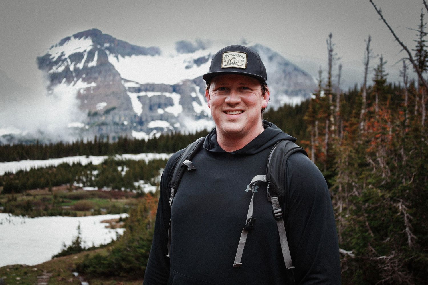 Smiling hiker wearing a black cap and backpack stands before a snow-capped mountain landscape on a cloudy day.