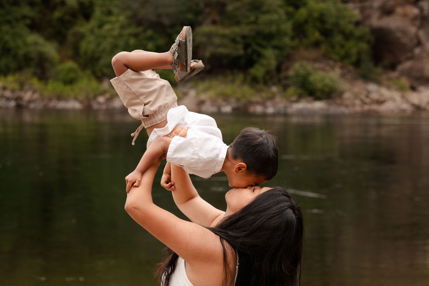A mother joyfully lifts her baby into the air near a serene lake surrounded by lush green trees.