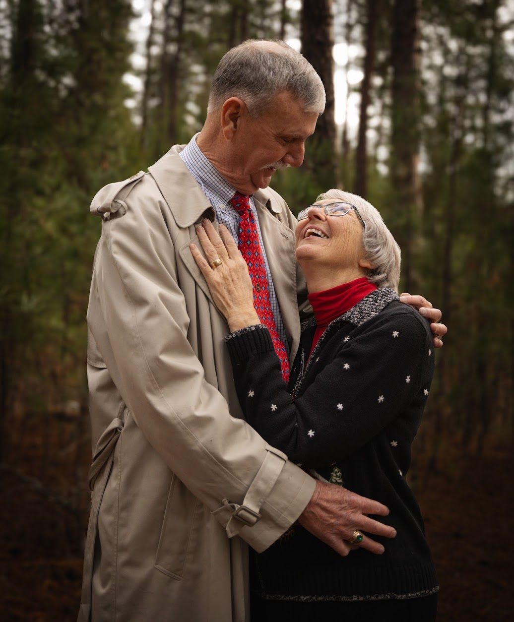 Elderly couple embracing and laughing together in a forest, man in trench coat and red tie, woman in dark sweater.