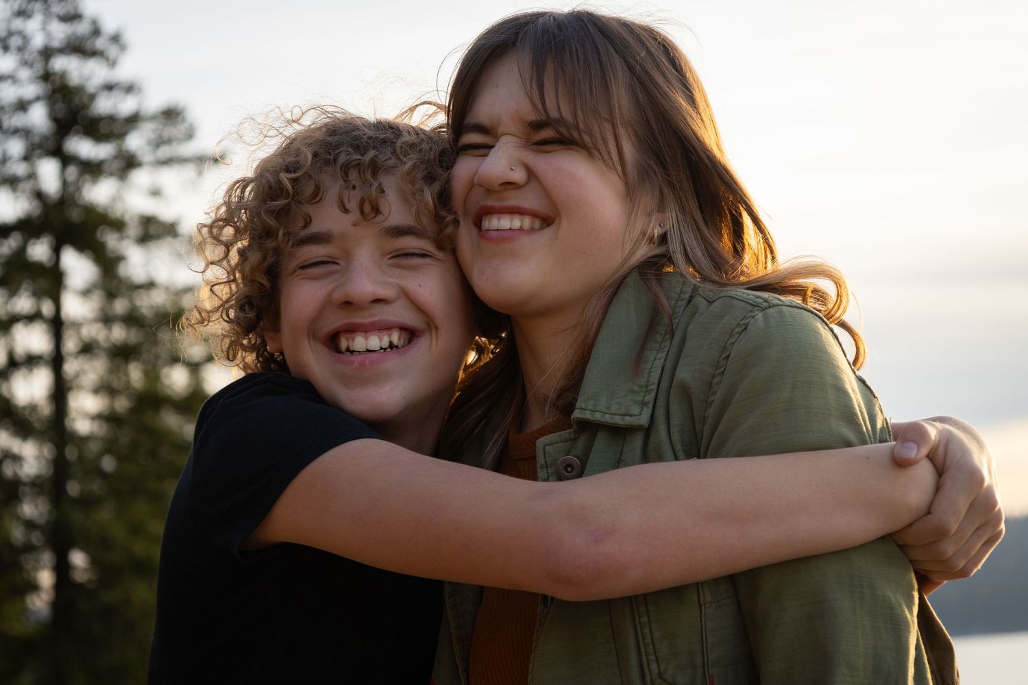 Two smiling teenagers hugging outdoors at golden hour with trees and water in the background.