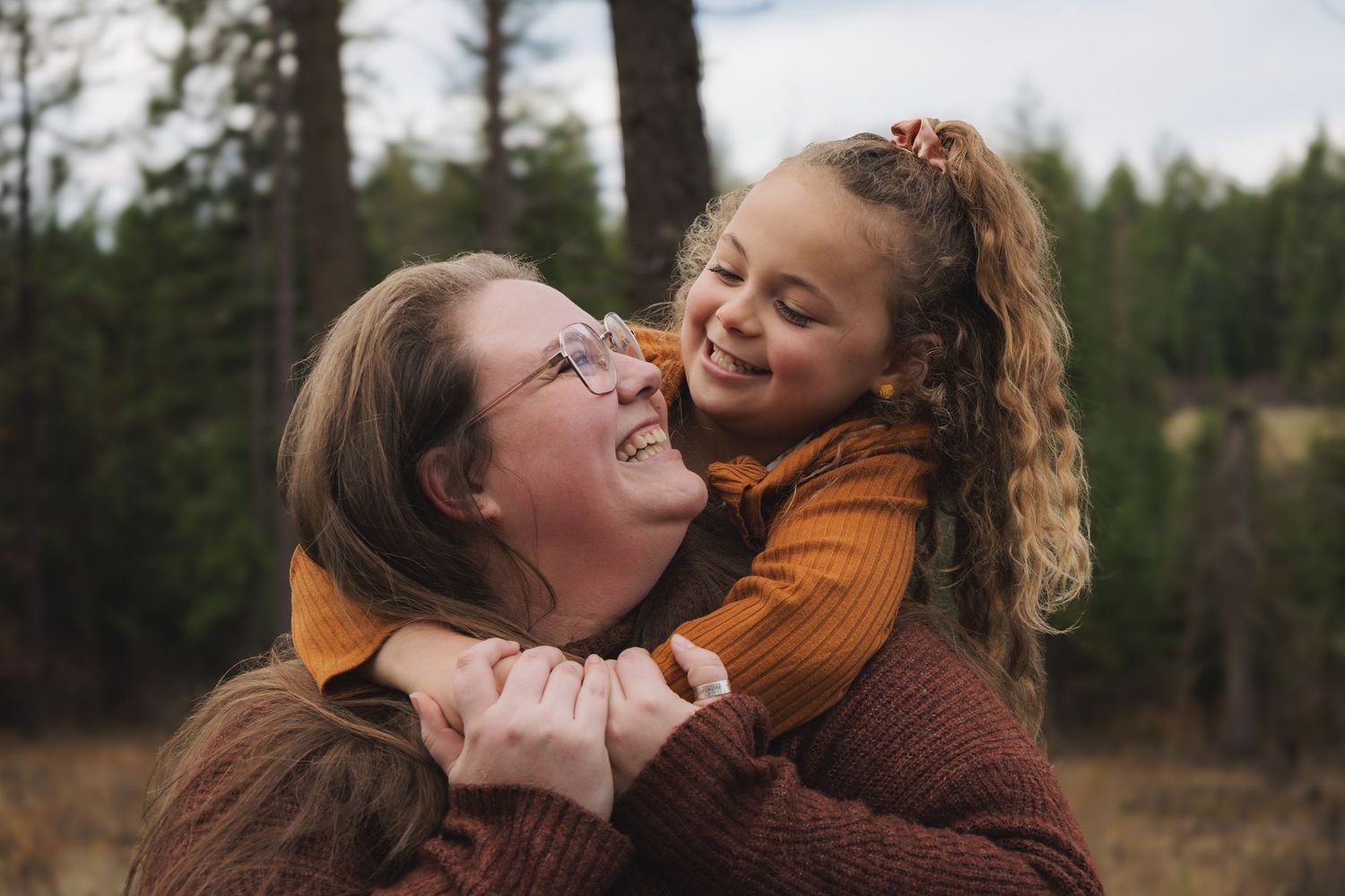 A mother joyfully hugs her smiling young daughter outdoors in a forest setting, both wearing warm autumn sweaters.