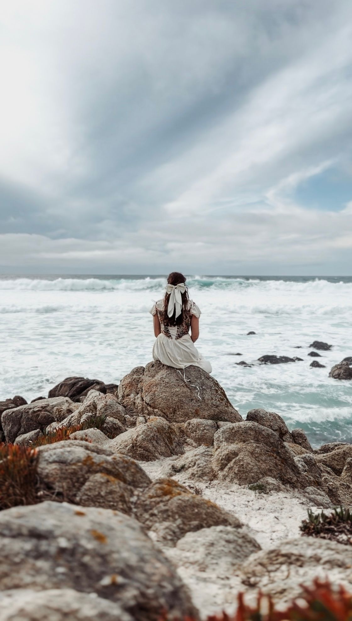 Woman in white dress sits on coastal rocks overlooking dramatic ocean waves under cloudy sky.