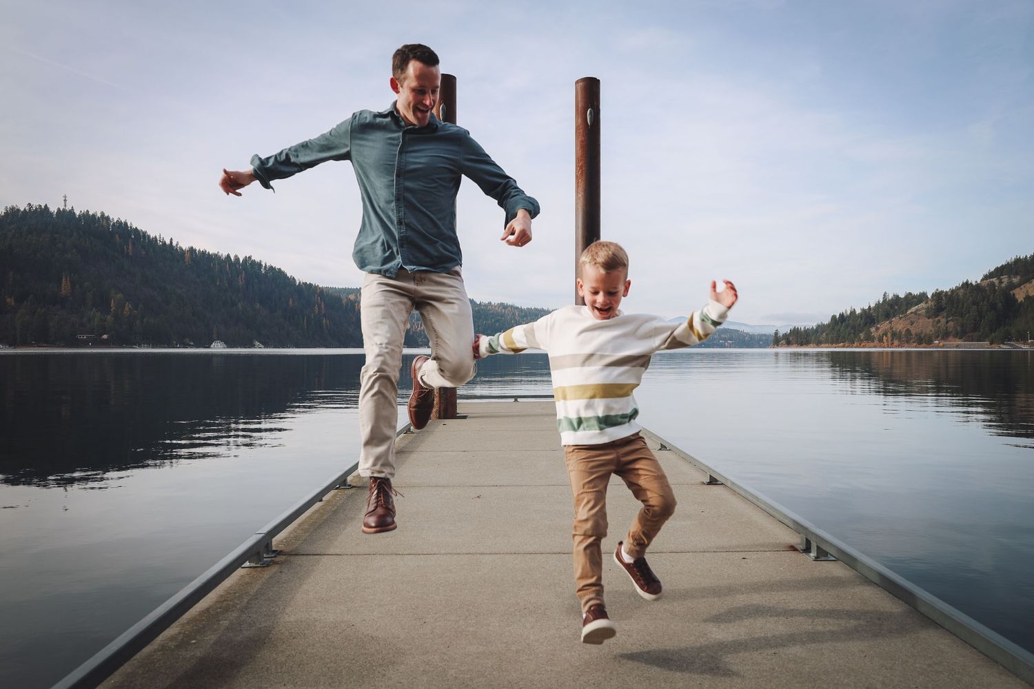 A father and son joyfully jump together on a wooden dock overlooking a calm lake.