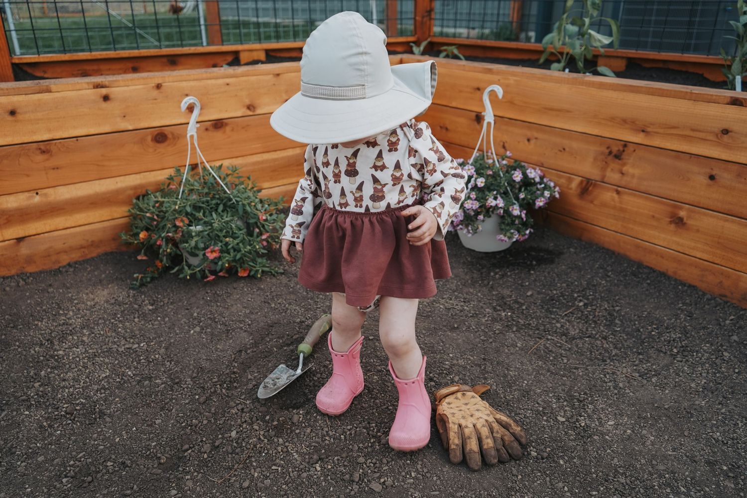 Toddler girl in sun hat and pink boots gardening with shovel and gloves in raised bed garden.