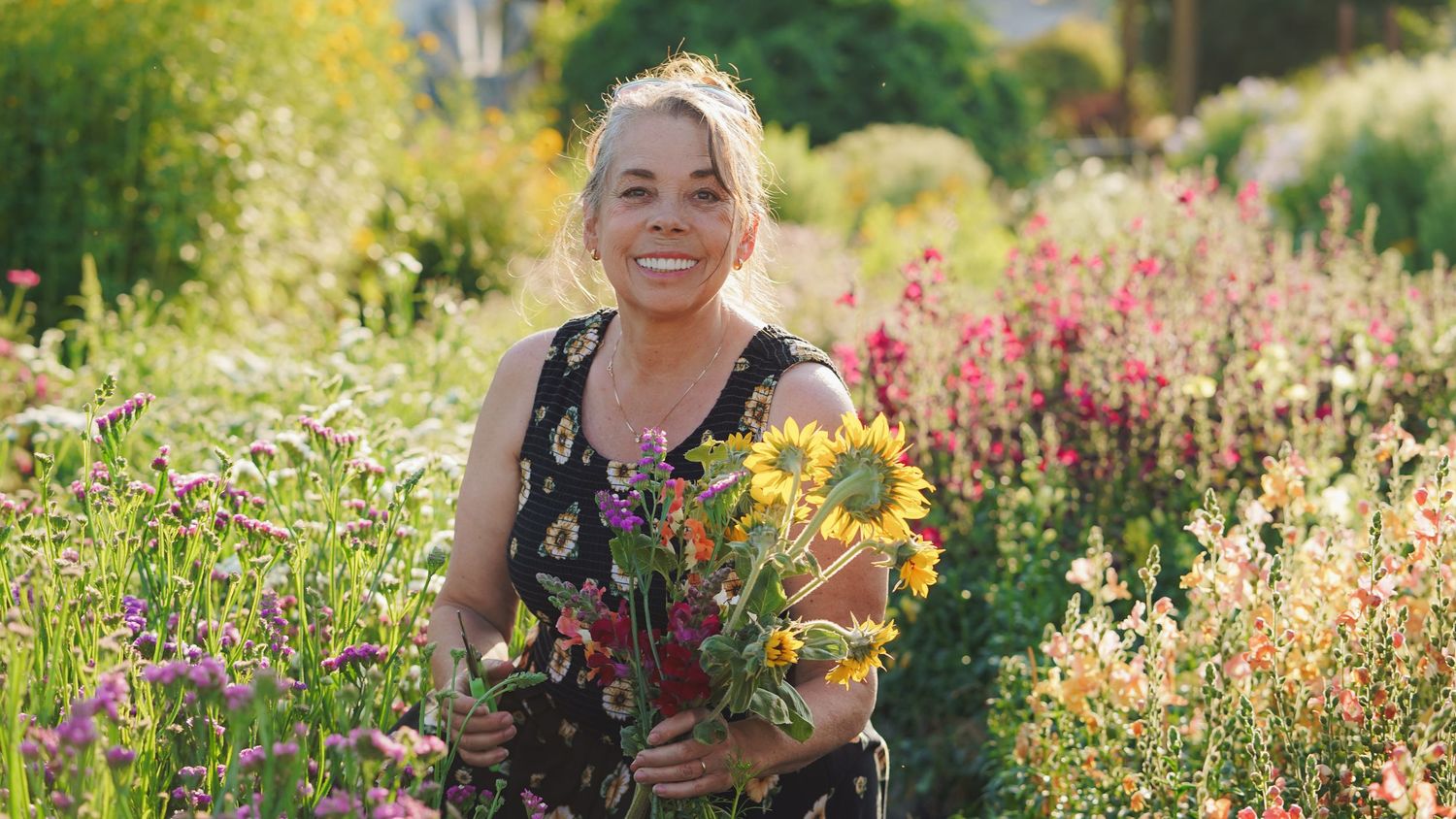 Woman smiling in a vibrant flower garden holding a colorful bouquet with sunflowers on a sunny day.