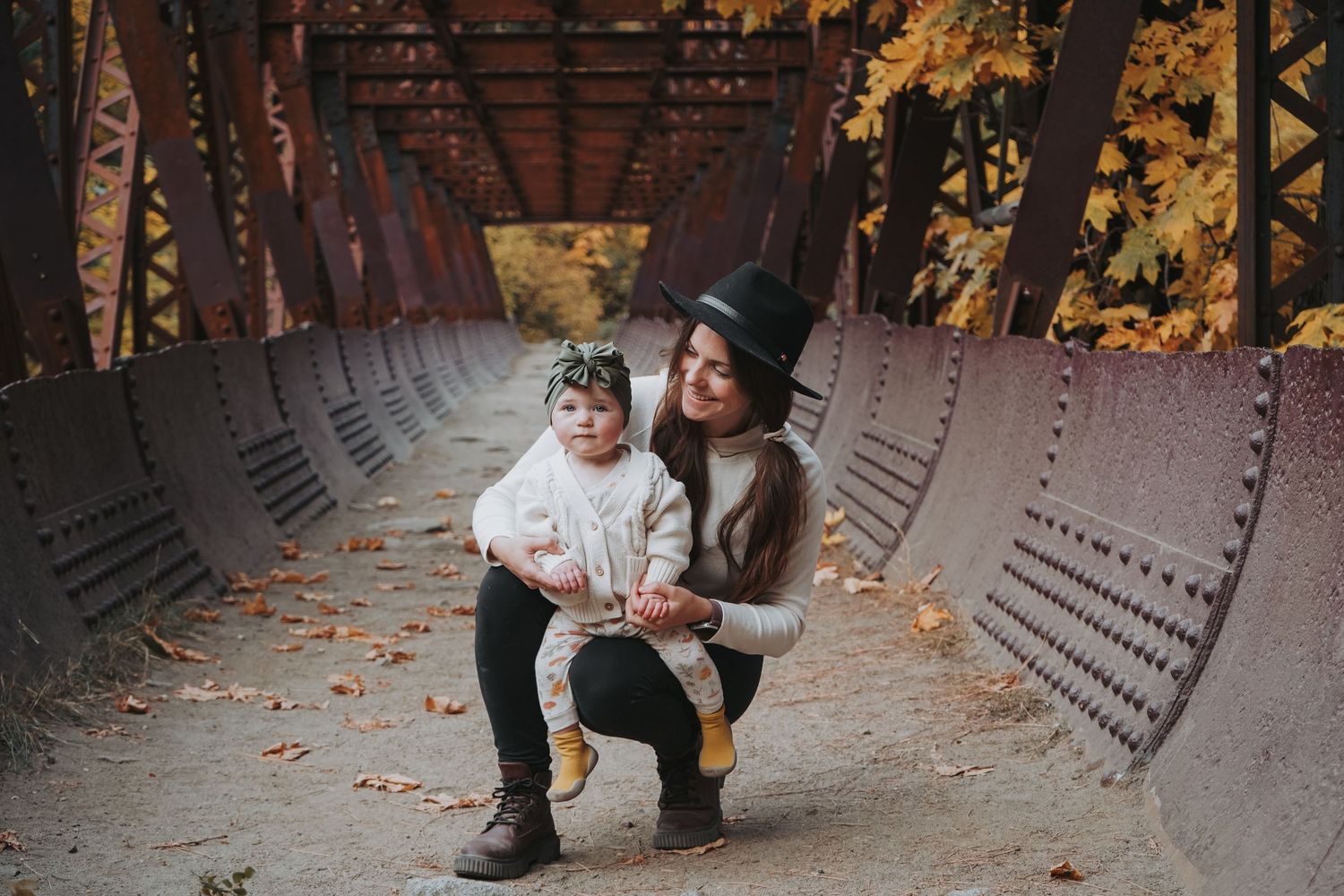 A mother crouches beside her flower-crowned toddler on a rustic autumn bridge.