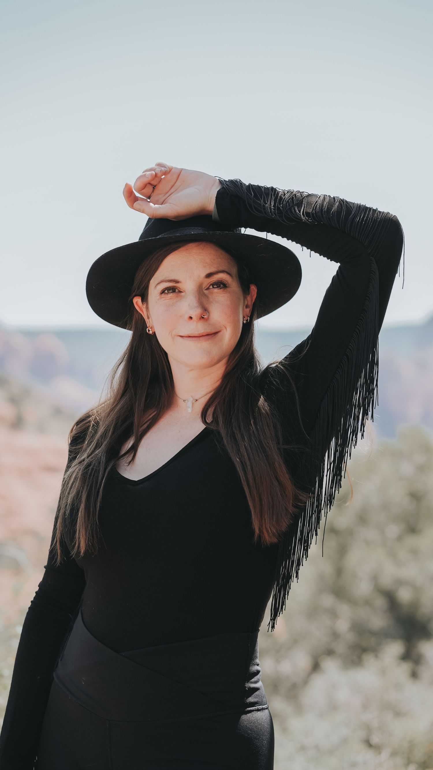 Stylish brunette woman in black fringe jacket and hat poses against desert canyon backdrop.
